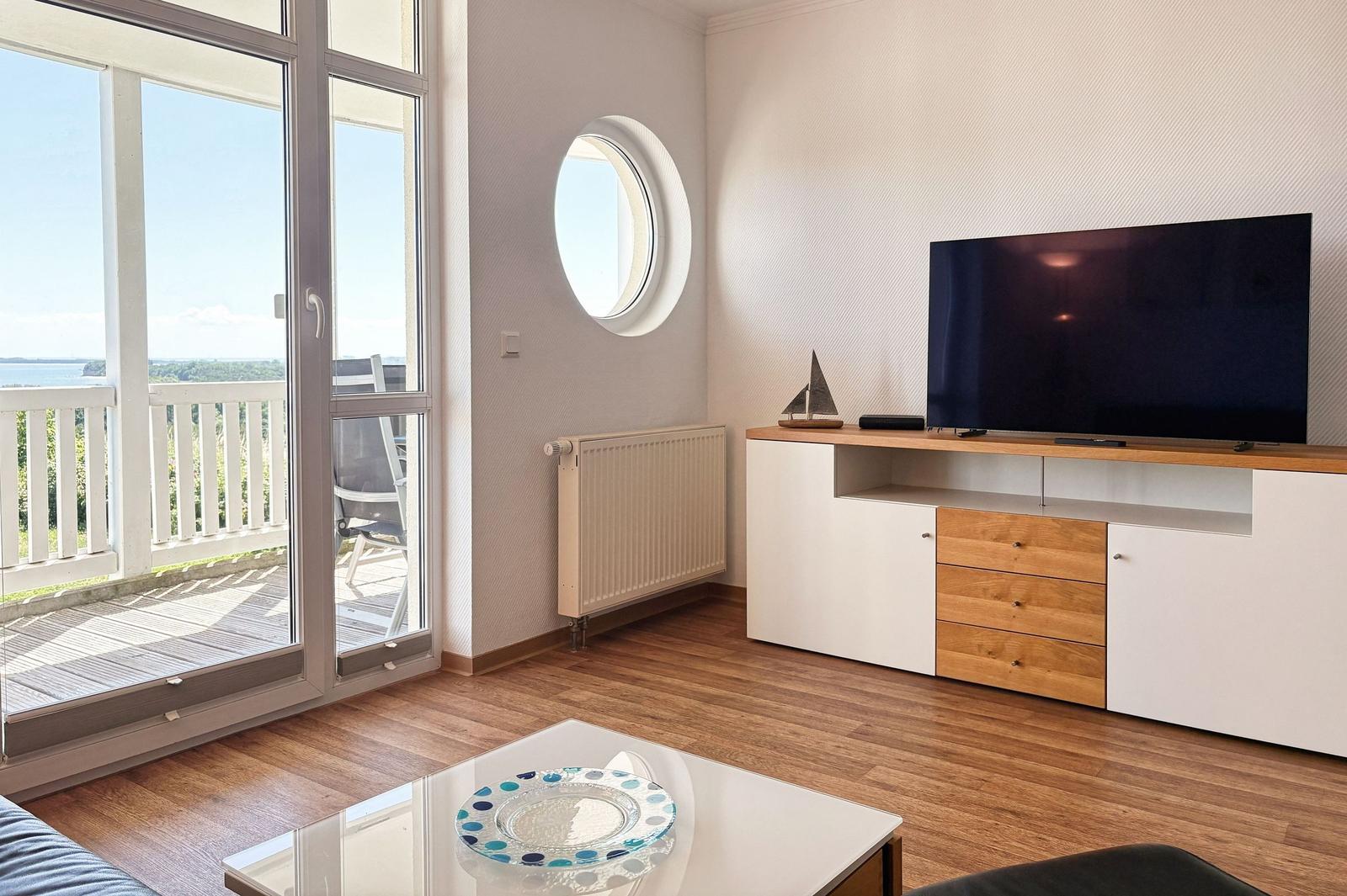 Wohnzimmer mit TV, Rundfenster und Balkonblick. Holzfußboden und moderner Schrank.