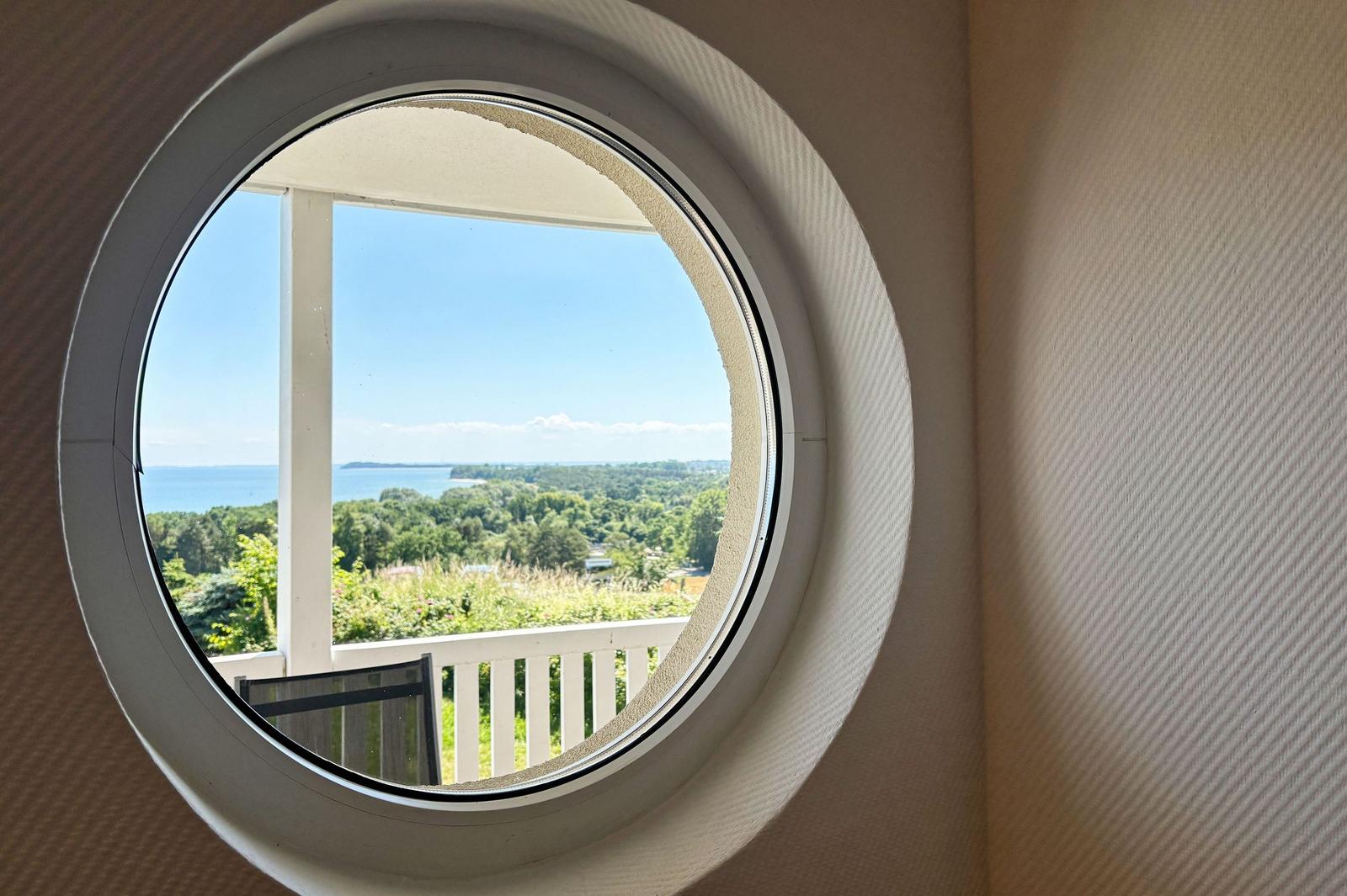 Rundes Fenster zeigt Blick auf Balkon, Grünflächen und Meer.