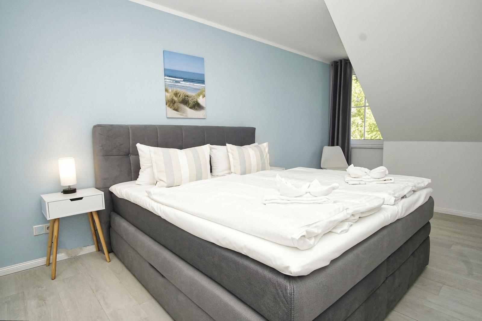 Schlafzimmer mit großem Bett, Nachttisch und Wandbild. Fenster mit Blick nach draußen.
