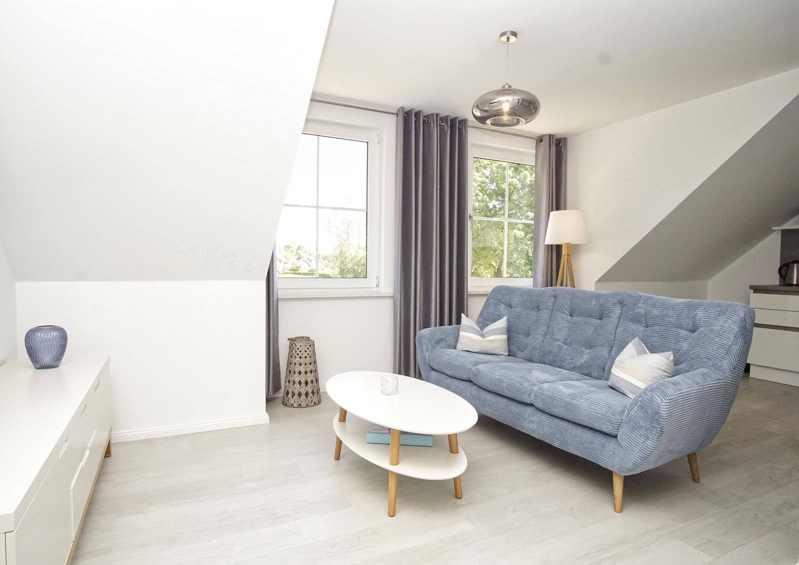 Wohnzimmer mit blauem Sofa, ovalen Tisch und Fenstern mit Vorhängen.