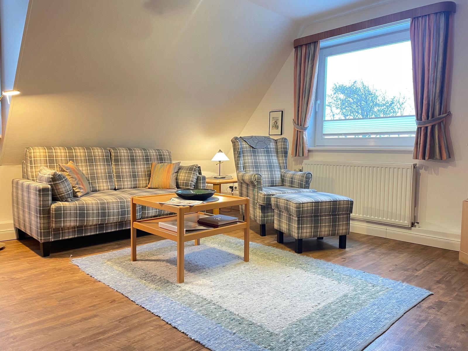 Wohnzimmer mit Sofa, Sessel und Tisch. Fenster mit Vorhängen und Holzboden.