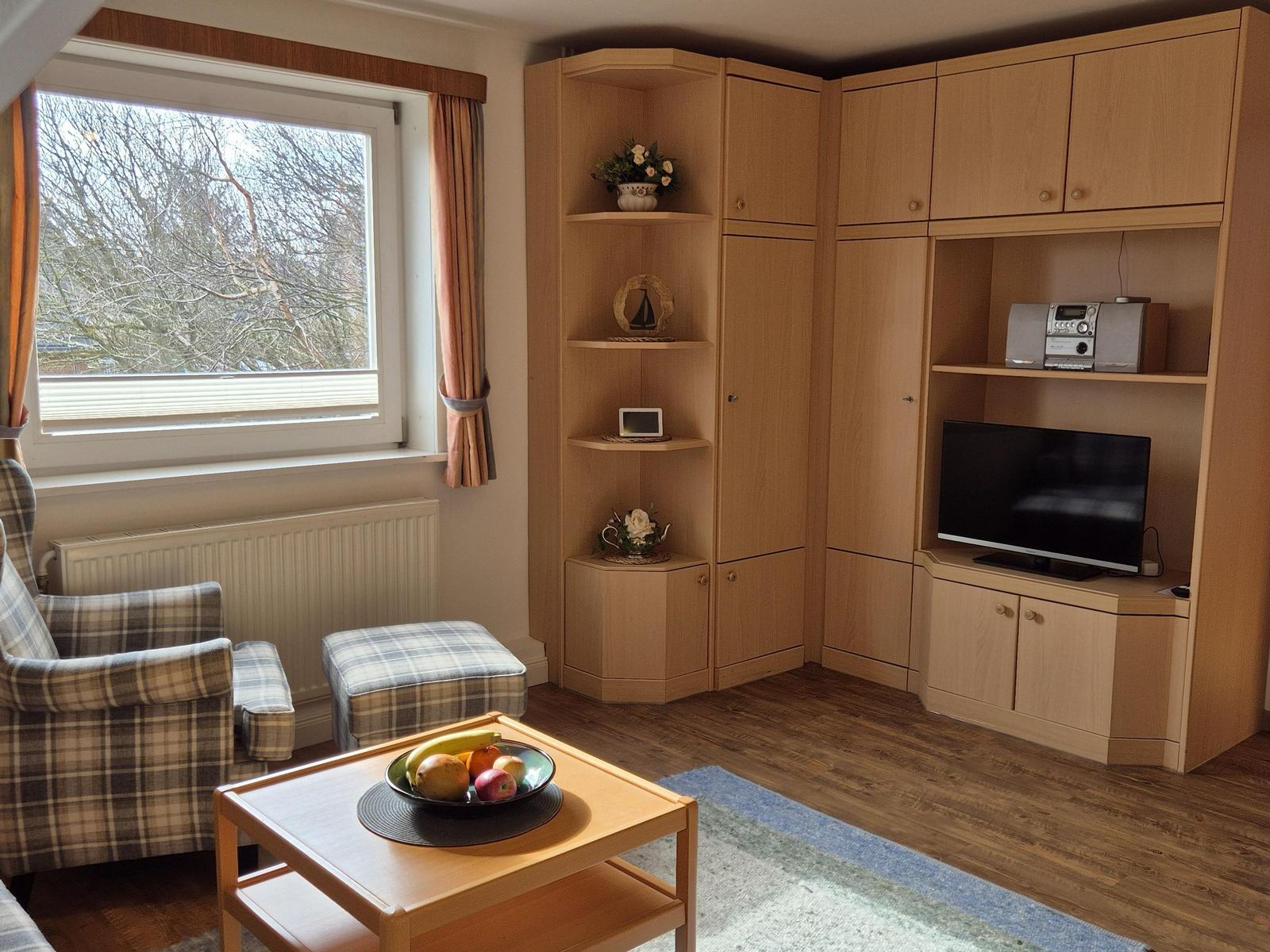 Wohnzimmer mit Holzmöbeln, Fernseher und Fenster mit Blick auf Bäume.
