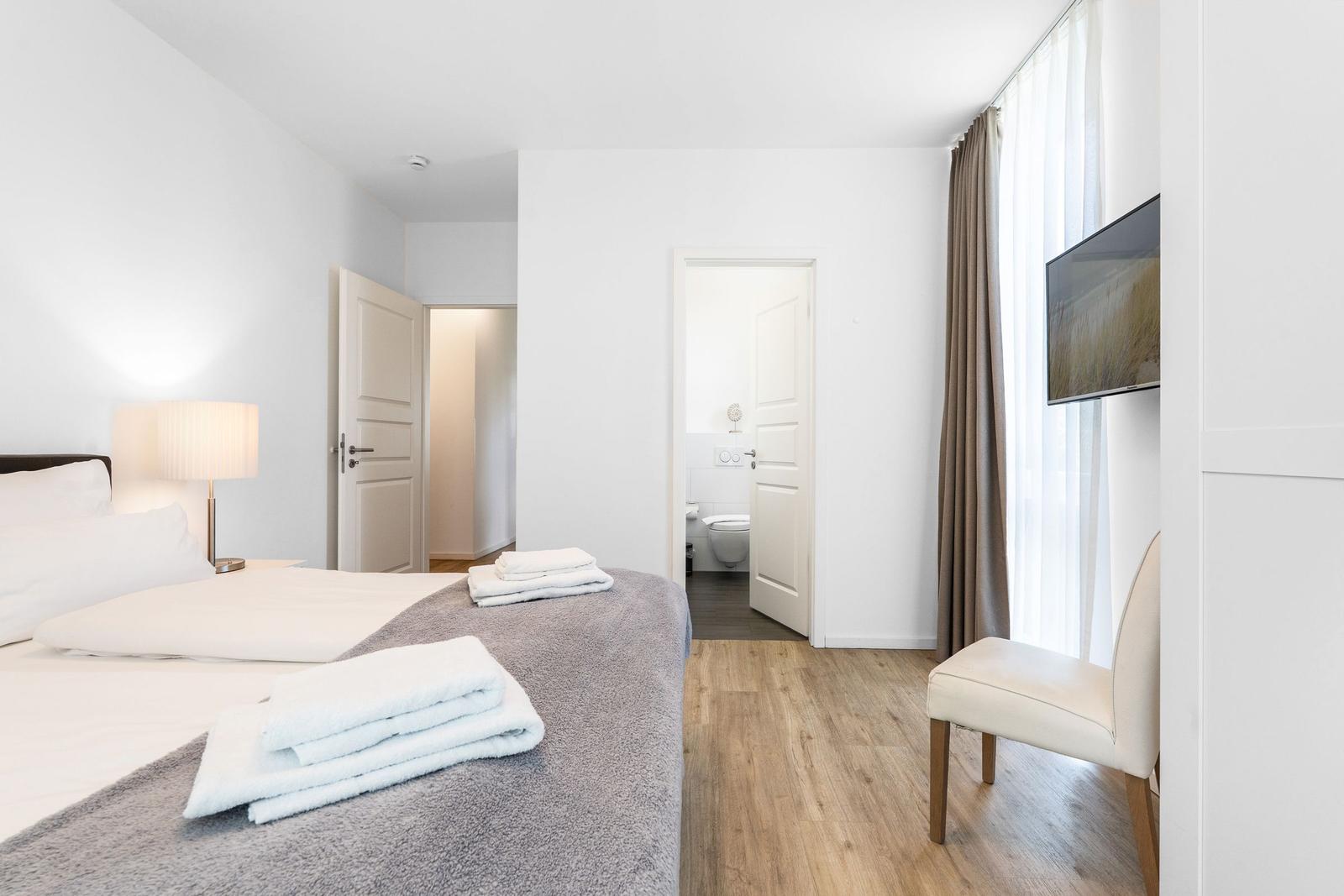 Schlafzimmer mit Bett, Fernseher und Bad. Holzfußboden und Fenster mit Vorhängen.