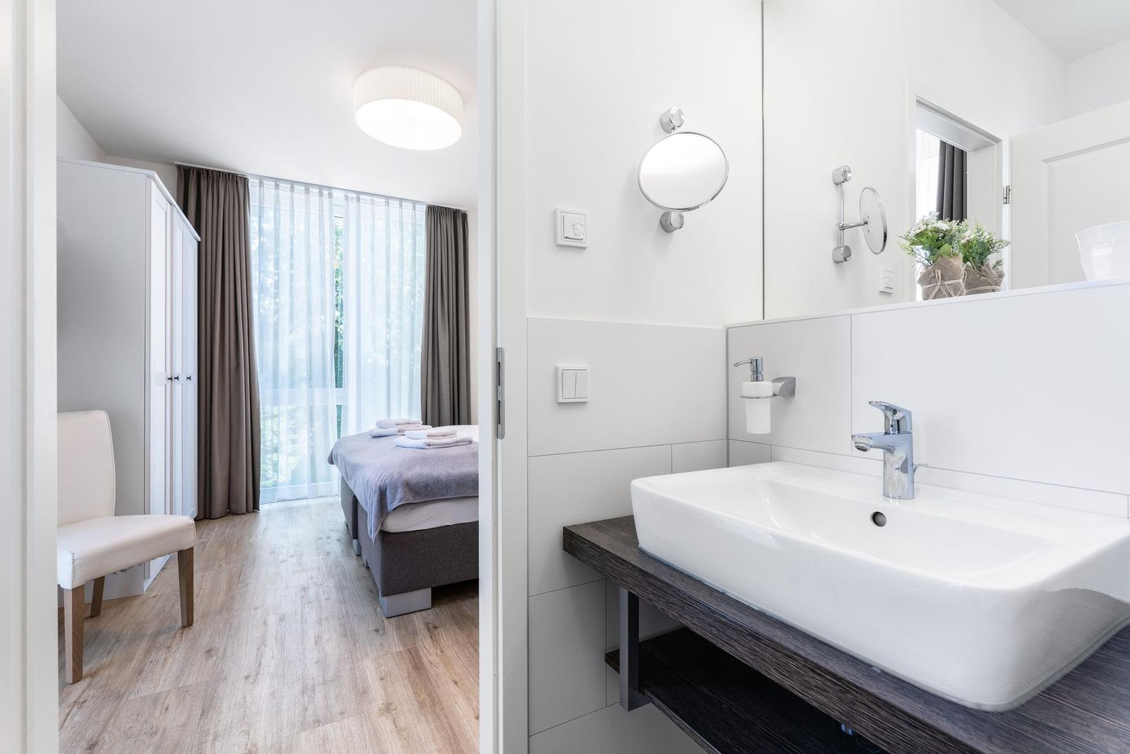 Badezimmer mit Waschbecken und Spiegel. Im Hintergrund ist ein Schlafzimmer mit Bett und Vorhang.