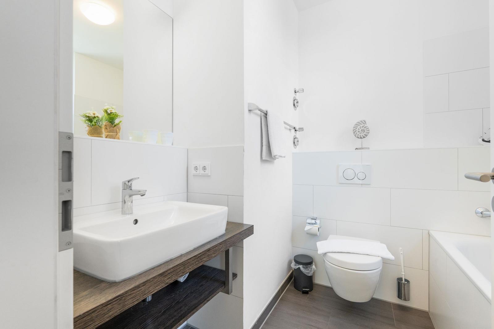 Badezimmer mit Waschbecken, Toilette und Badewanne. Weiße Fliesen und Holzunterschrank.