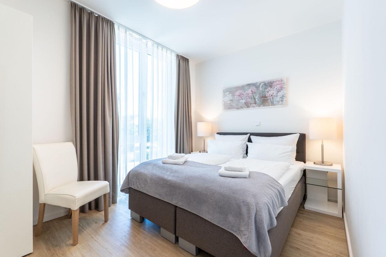 Schlafzimmer mit Doppelbett, zwei Nachttischen und einem Stuhl neben dem Fenster.