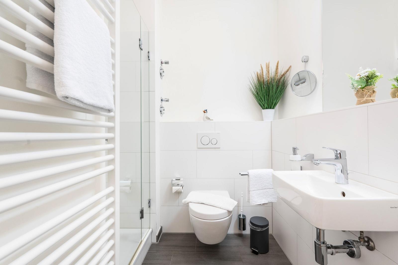 Badezimmer mit Dusche, Toilette und Waschbecken. Weiße Fliesen und Handtuchhalter.