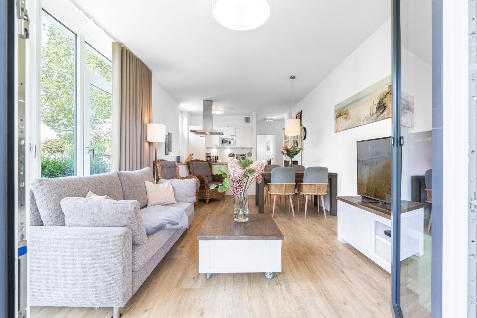 Wohnzimmer mit Sofa, Esstisch und TV. Holzboden und große Fenster.
