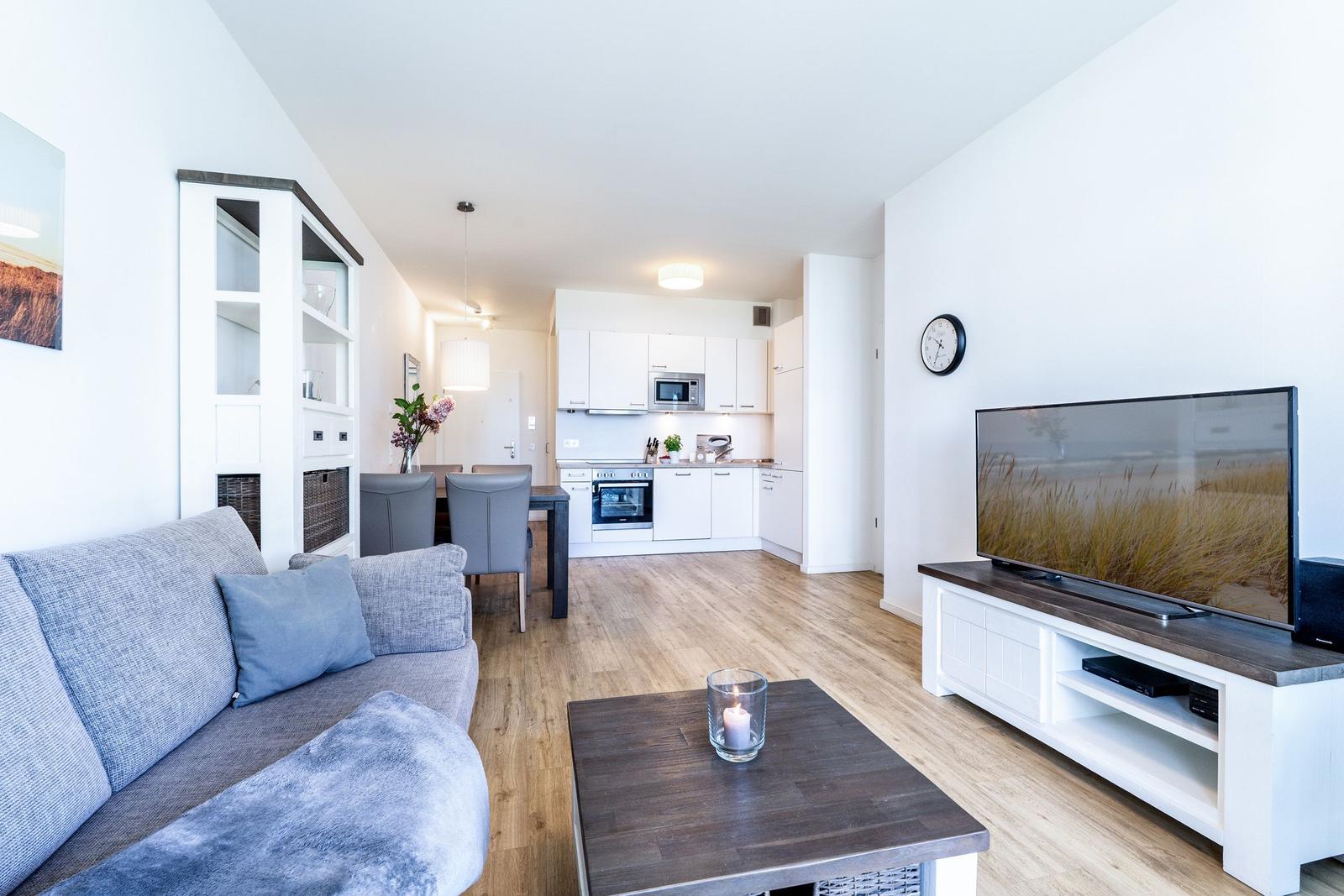 Wohnzimmer mit Sofa, Couchtisch und Fernseher. Küche im Hintergrund. Holzboden und weiße Wände.