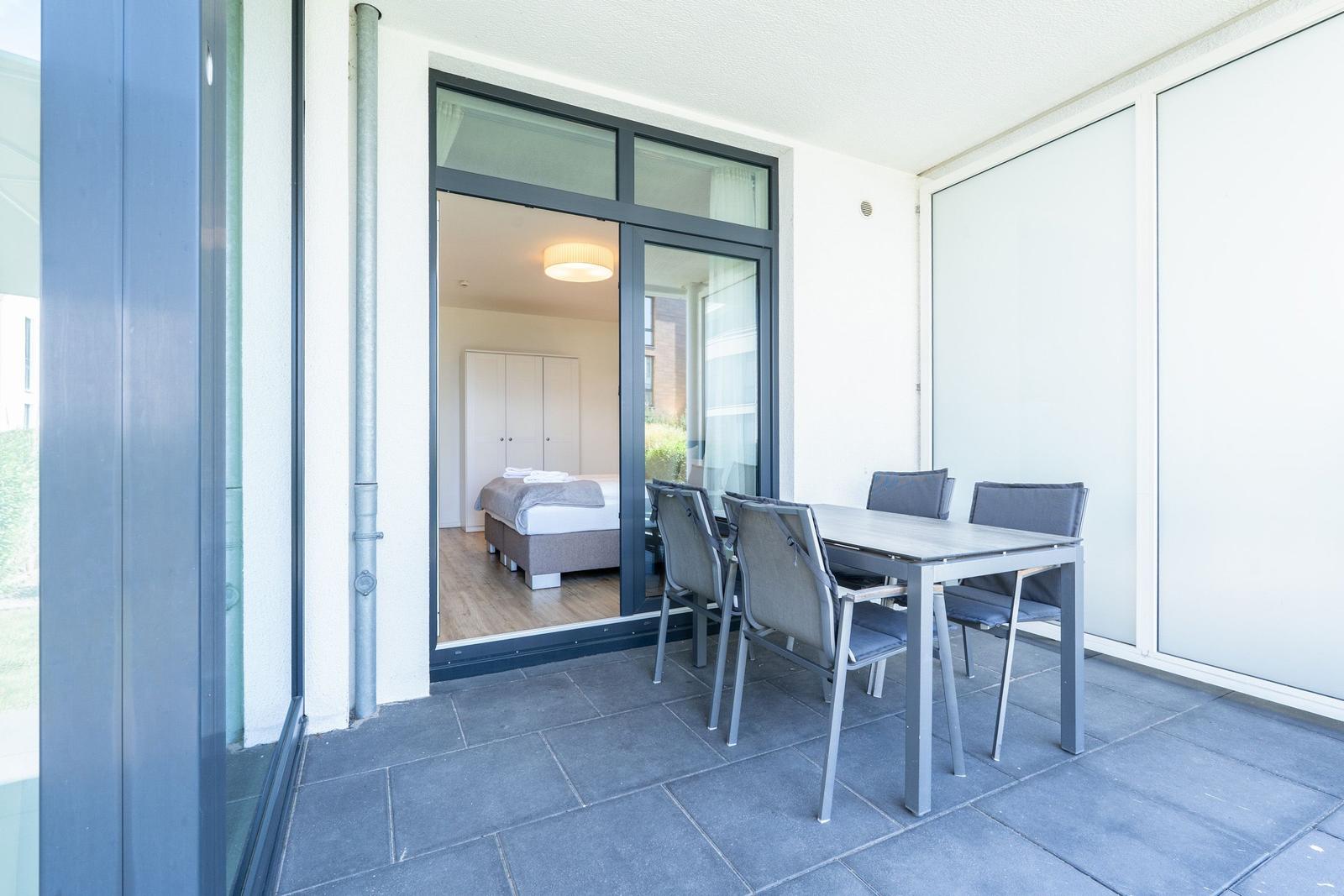 Terrasse mit Tisch und Stühlen. Durch Glas-Tür ist Schlafzimmer mit Bett sichtbar.