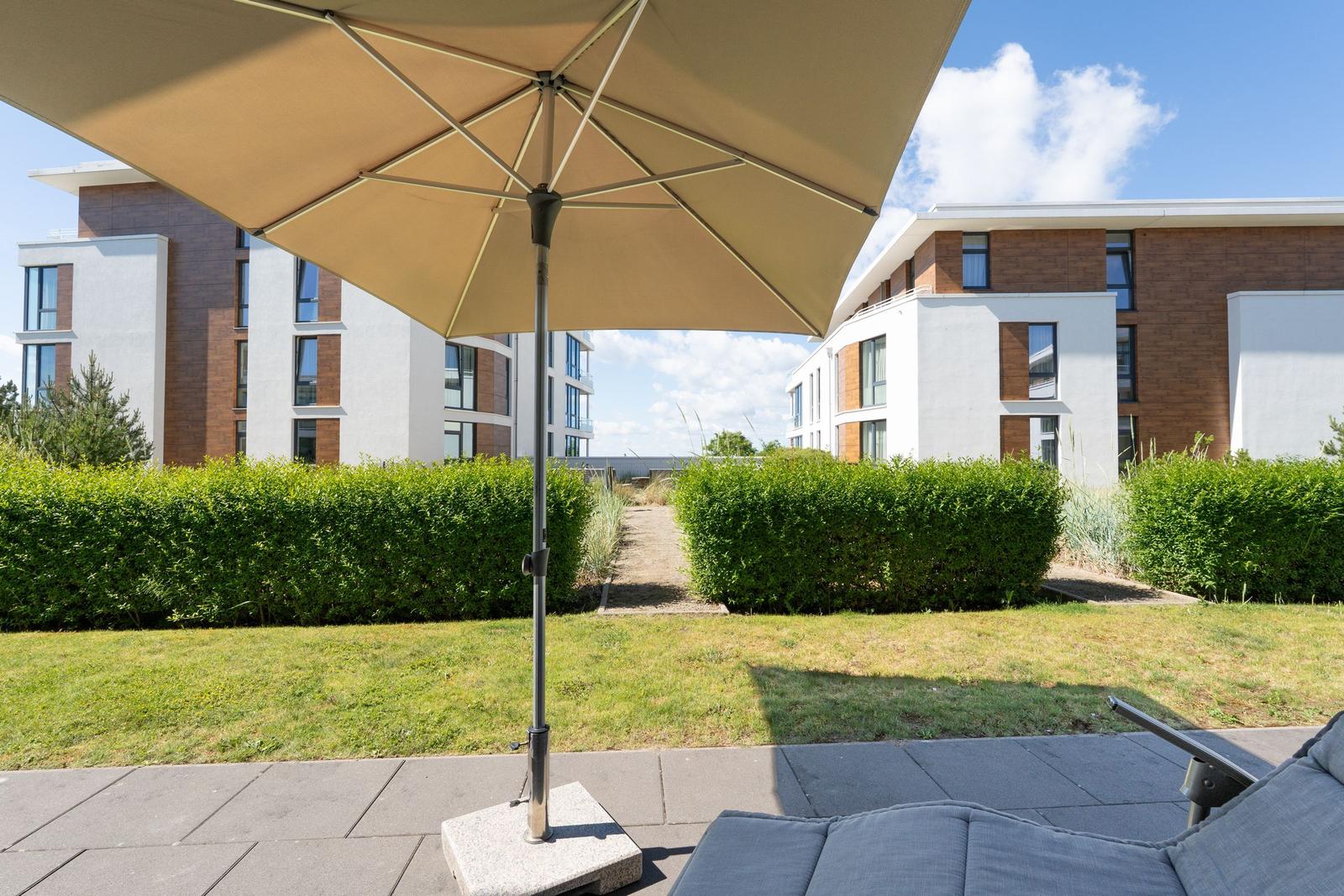 Terrasse mit Sonnenschirm und Liegefläche. Grünflächen und moderne Gebäude im Hintergrund.