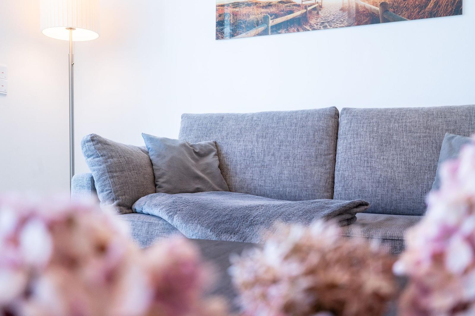 Ein grauer Sofa mit Kissen und einer Decke. Im Hintergrund eine Wandlampe und ein Bild.