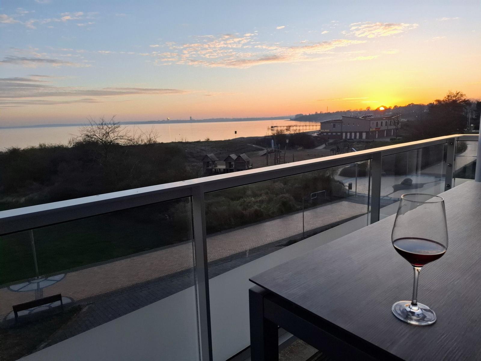 Ein Weinglas auf einer Terrasse mit Blick auf Sonnenuntergang über Wasser und Uferbereich.