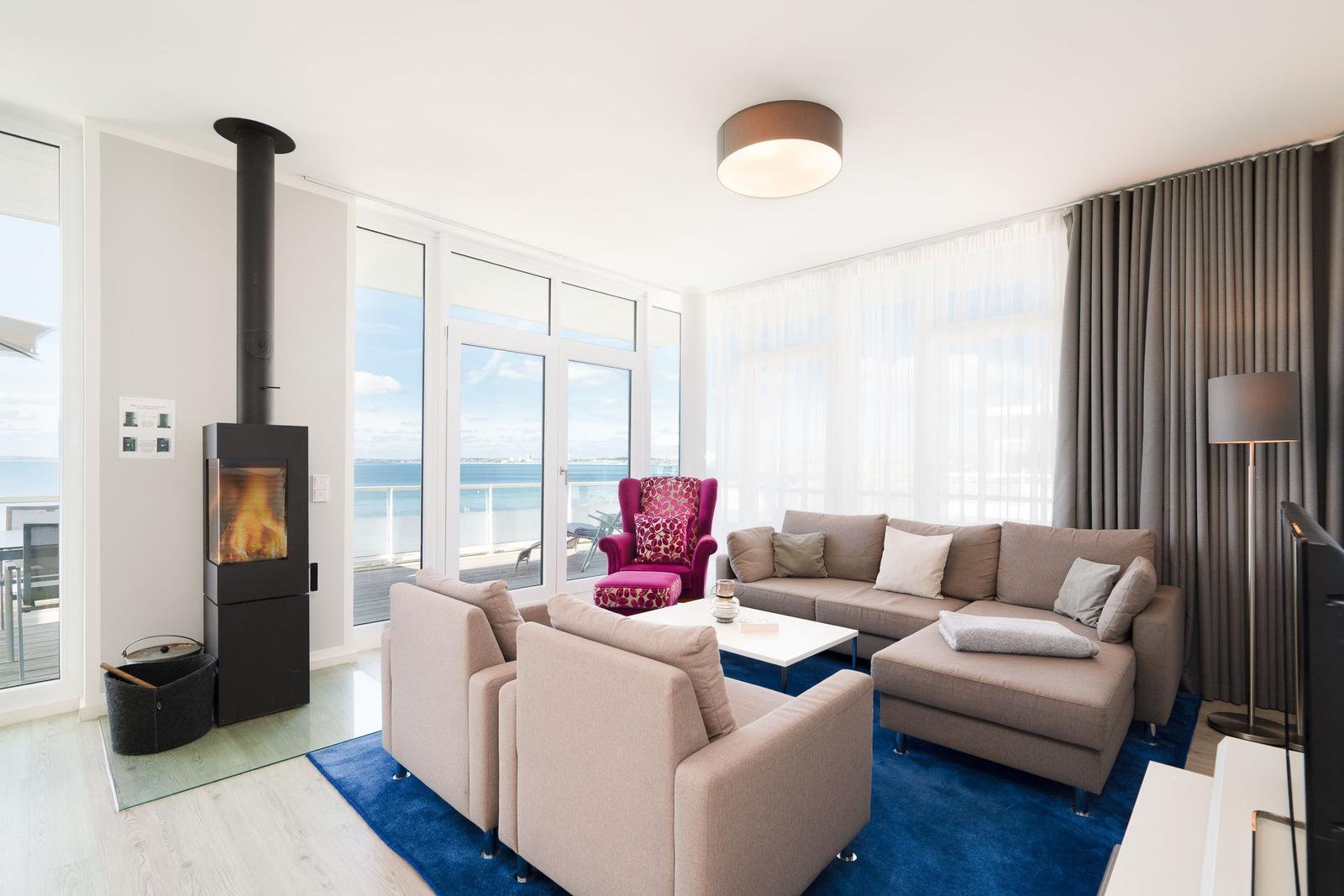 Wohnzimmer mit Sofa, Sessel und Kamin. Große Fenster mit Meerblick.