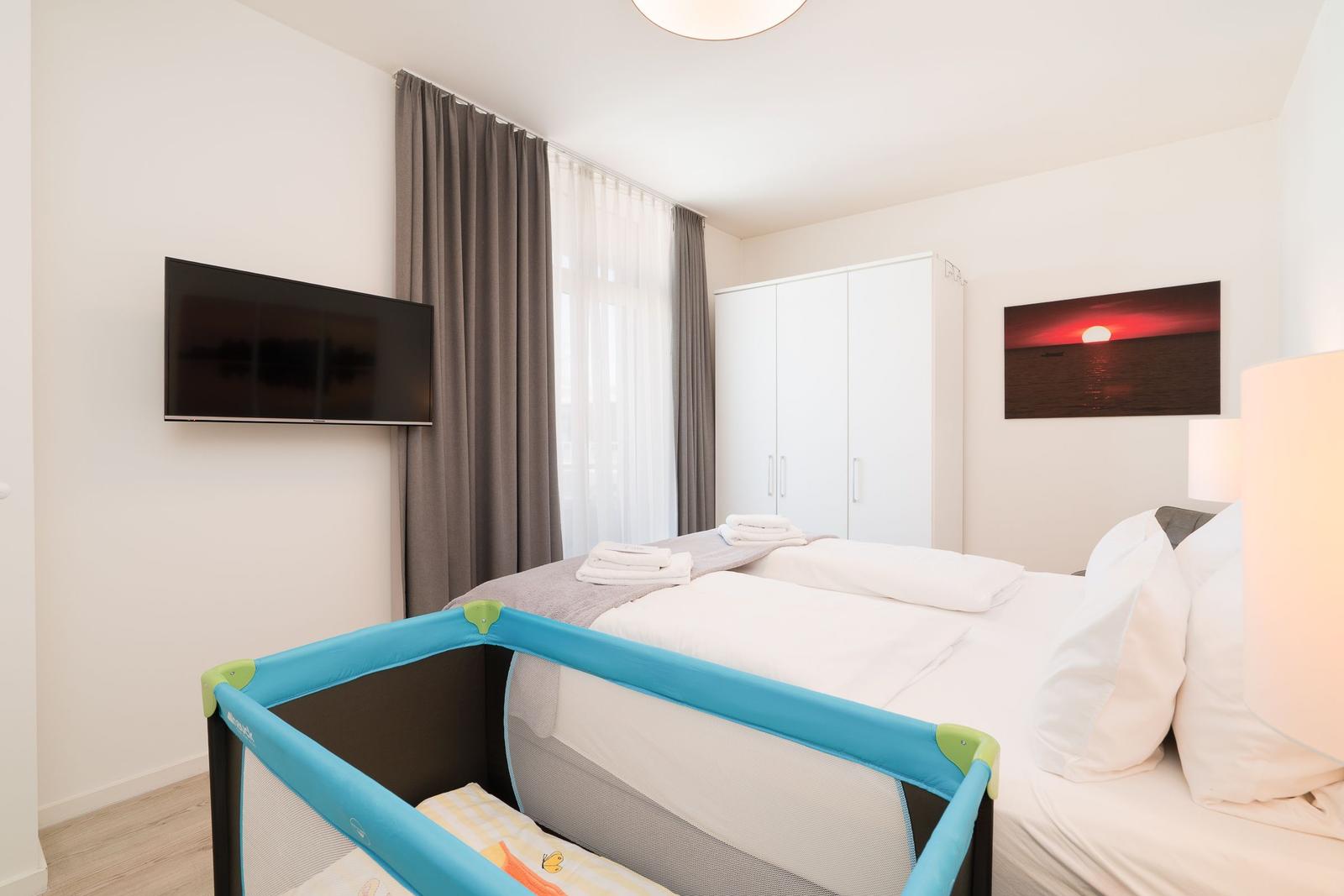 Schlafzimmer mit Bett, Kinderbett, TV und Kleiderschrank. Fenster mit Vorhängen.