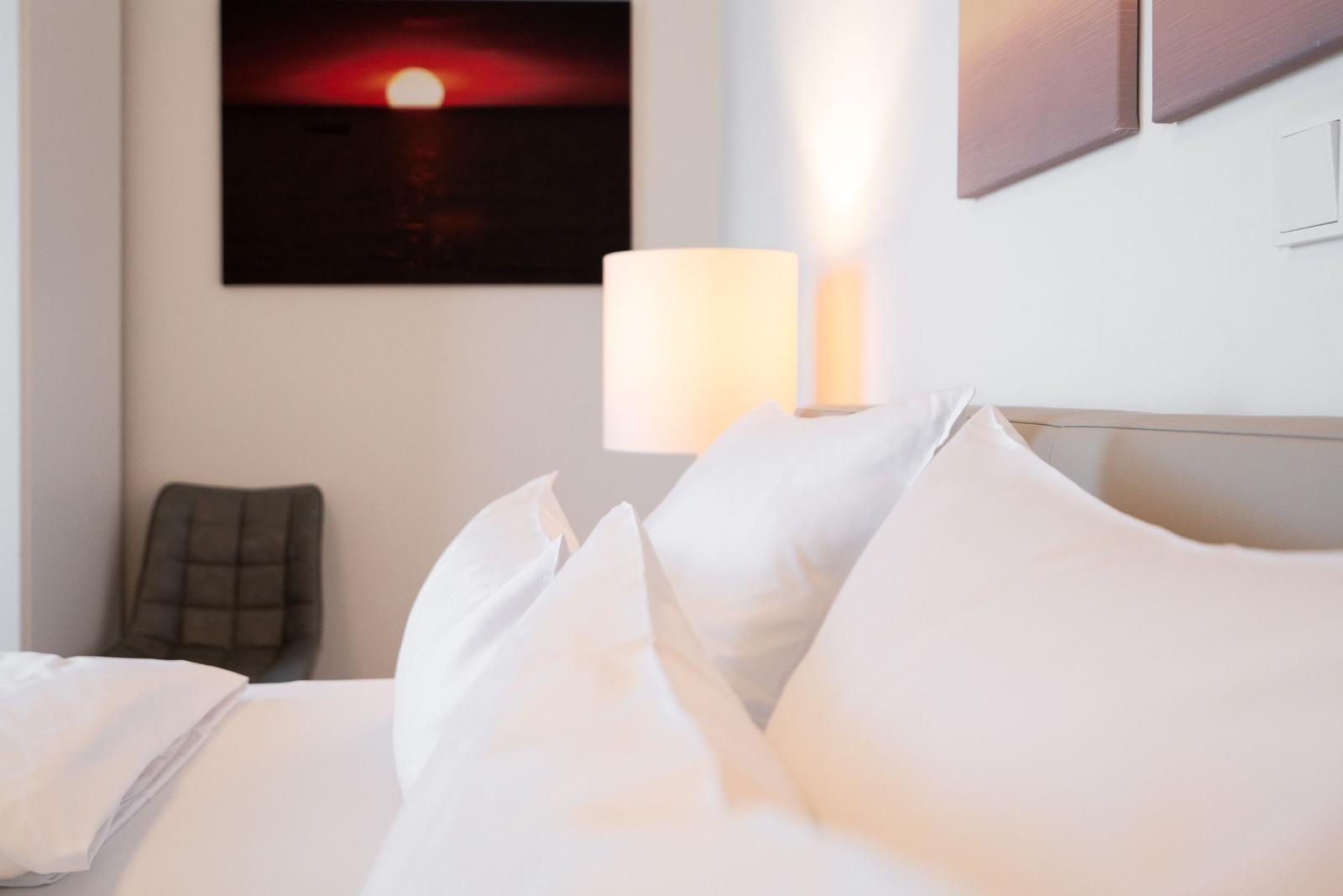 Bettkopf mit weißen Kissen, Nachttischlampe und Wandbild mit Sonnenuntergang.