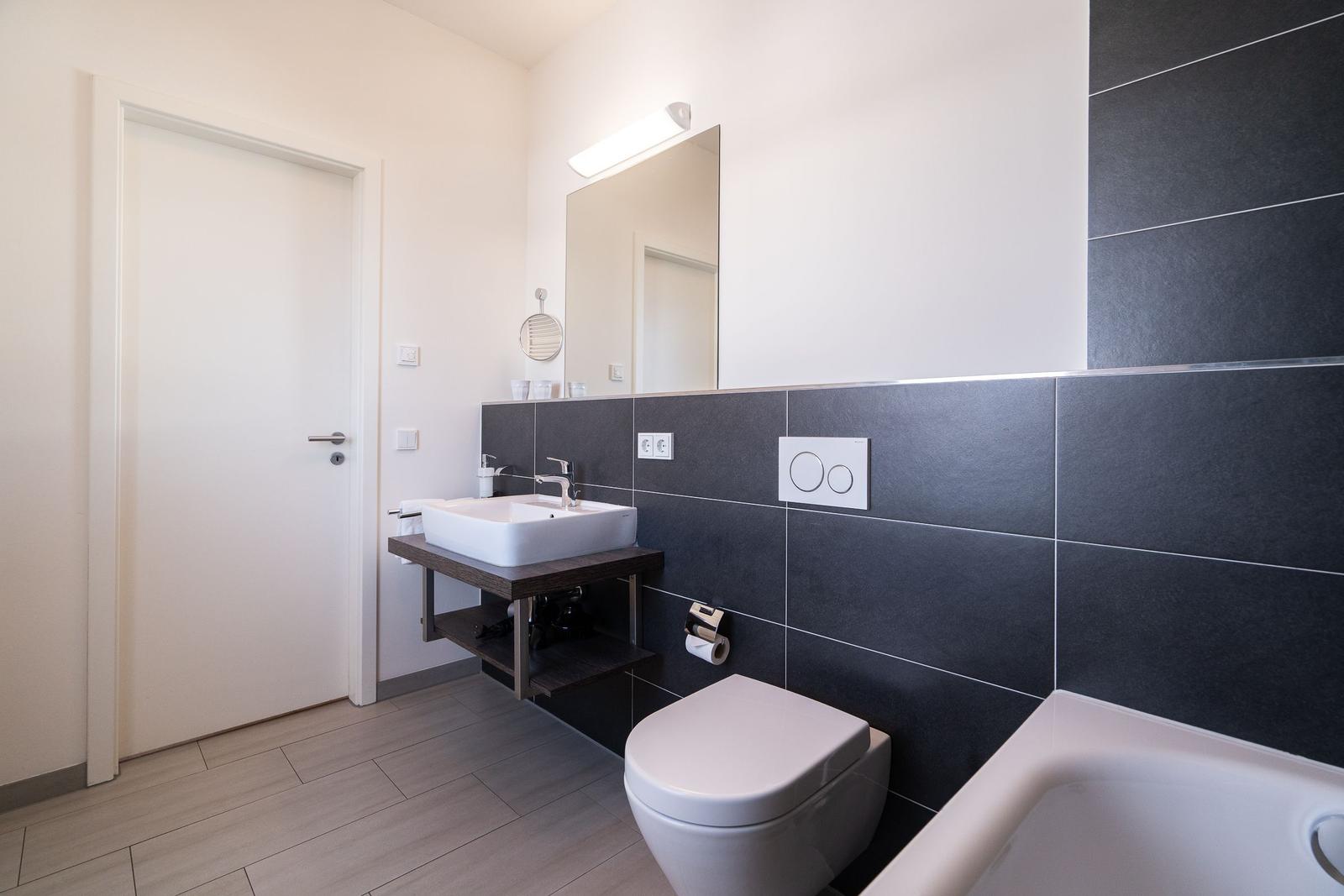 Badezimmer mit Waschbecken, Toilette und Badewanne. Schwarze Wandfliesen und Holzunterschrank.