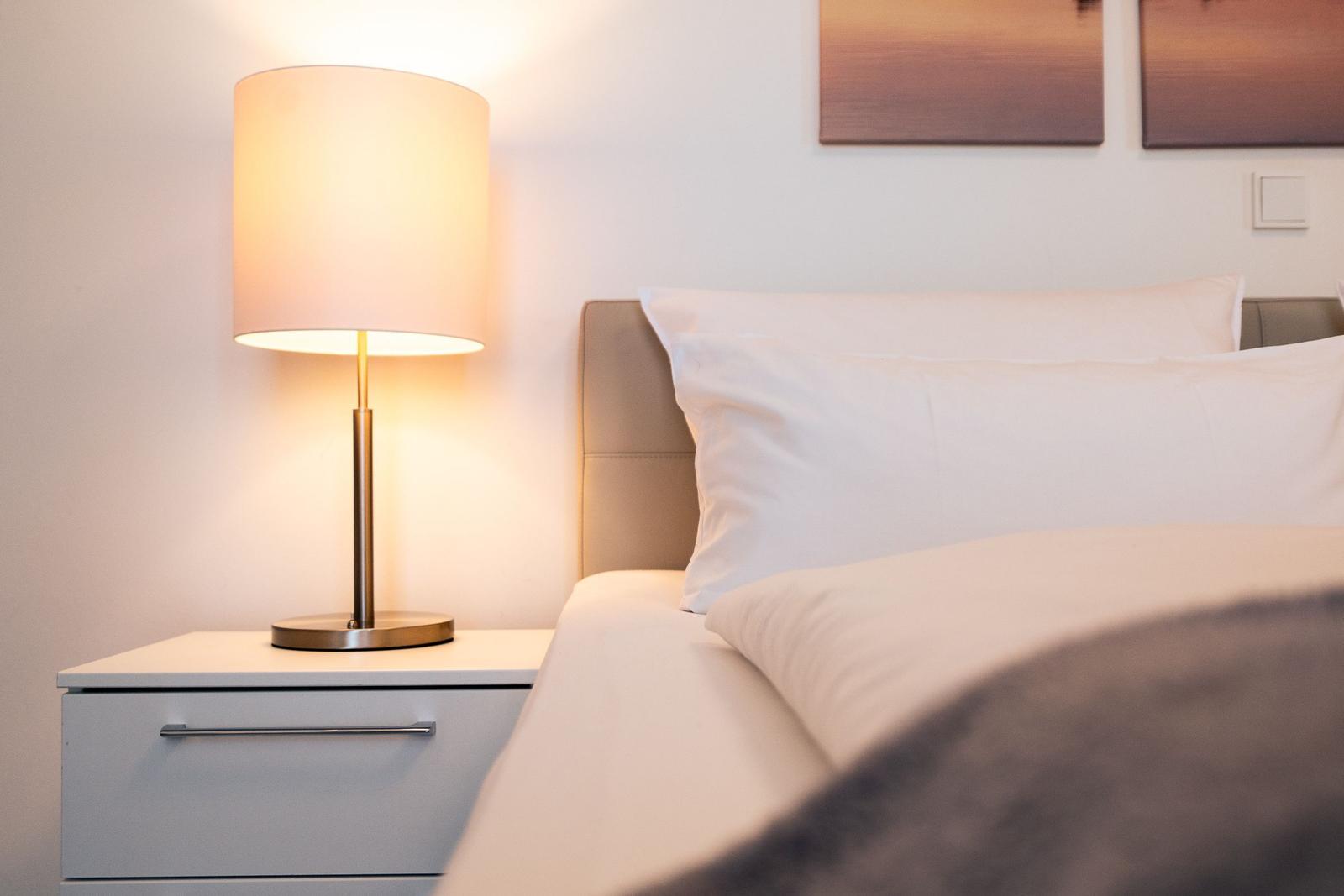 Bettkopfteil mit weißen Kissen, Nachttisch mit Lampe und Wandbildern.