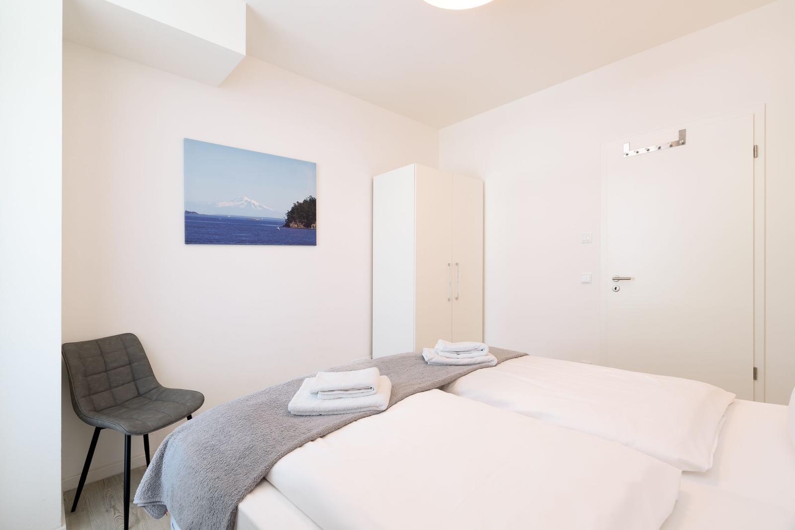 Schlafzimmer mit Bett, Kleiderschrank und Stuhl. Wandbild zeigt Berg und Meer.