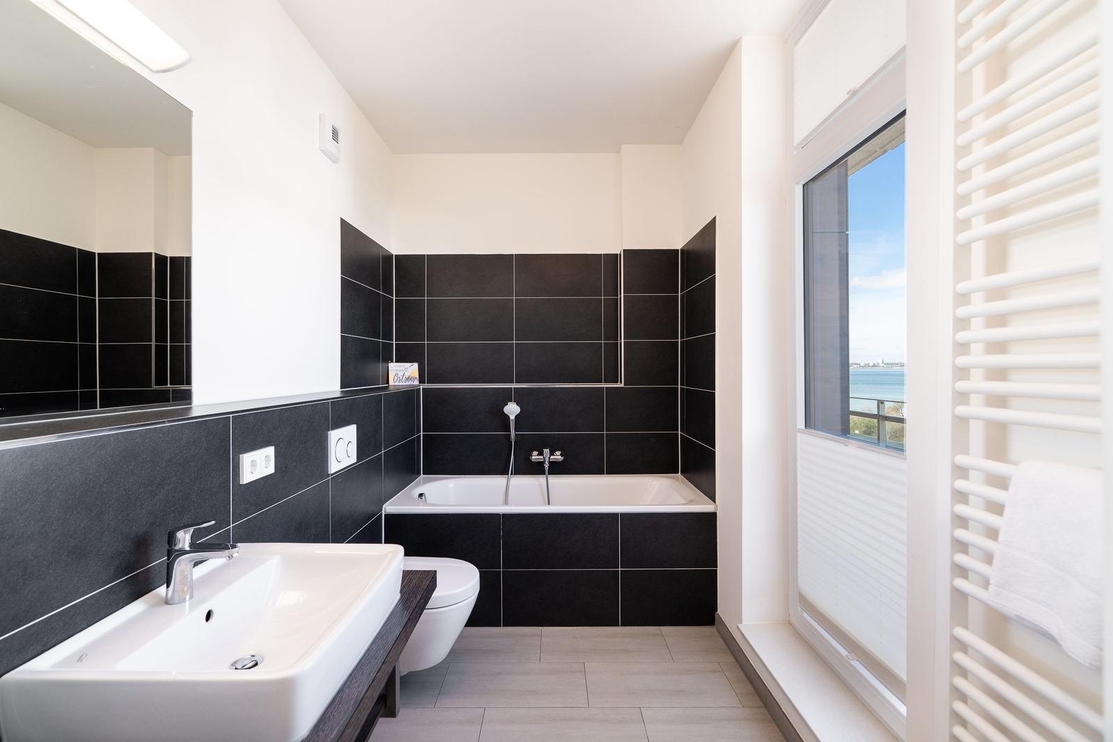 Badezimmer mit Badewanne, Waschbecken und Fensterblick auf Wasser.