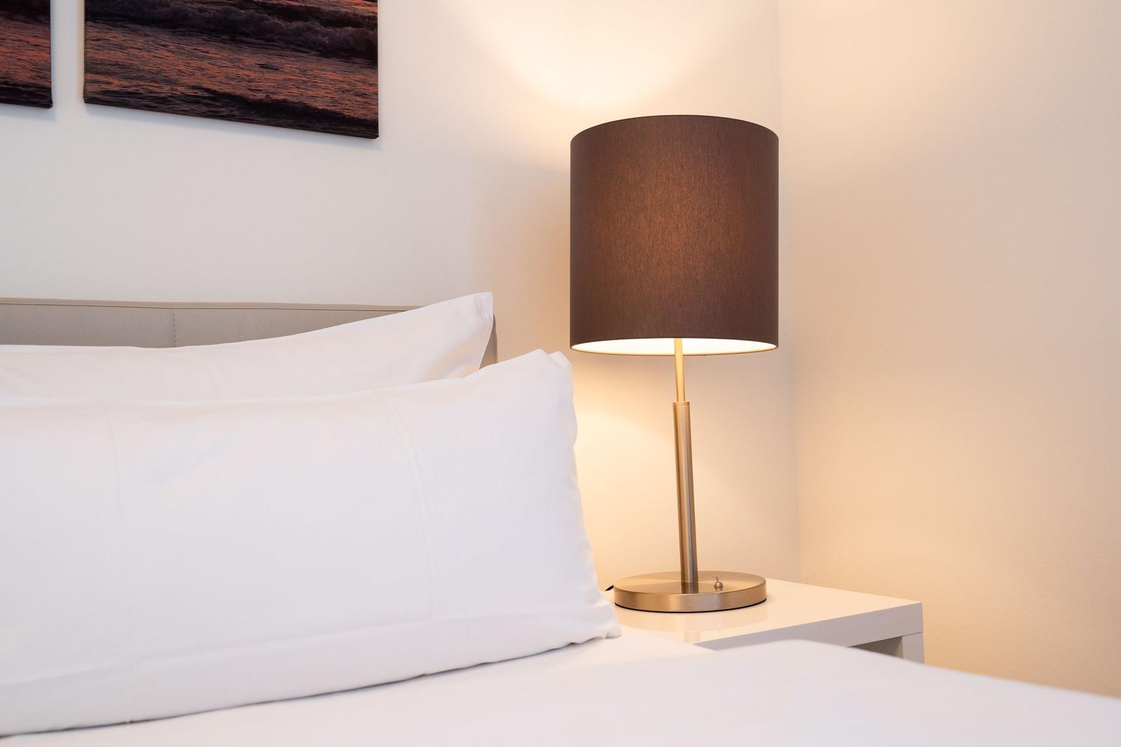 Schlafzimmer mit weißem Bett, Nachttisch und Stehlampe mit braunem Schirm.