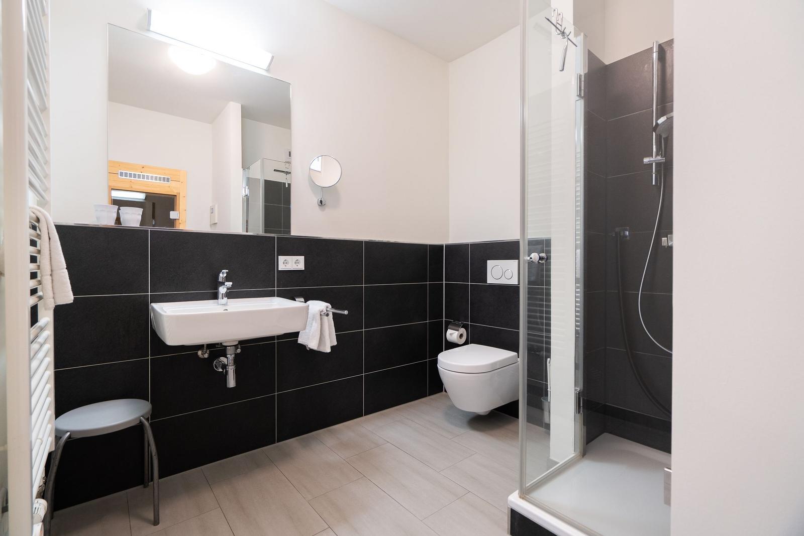 Badezimmer mit Dusche, Toilette und Waschbecken an schwarzen Fliesen.