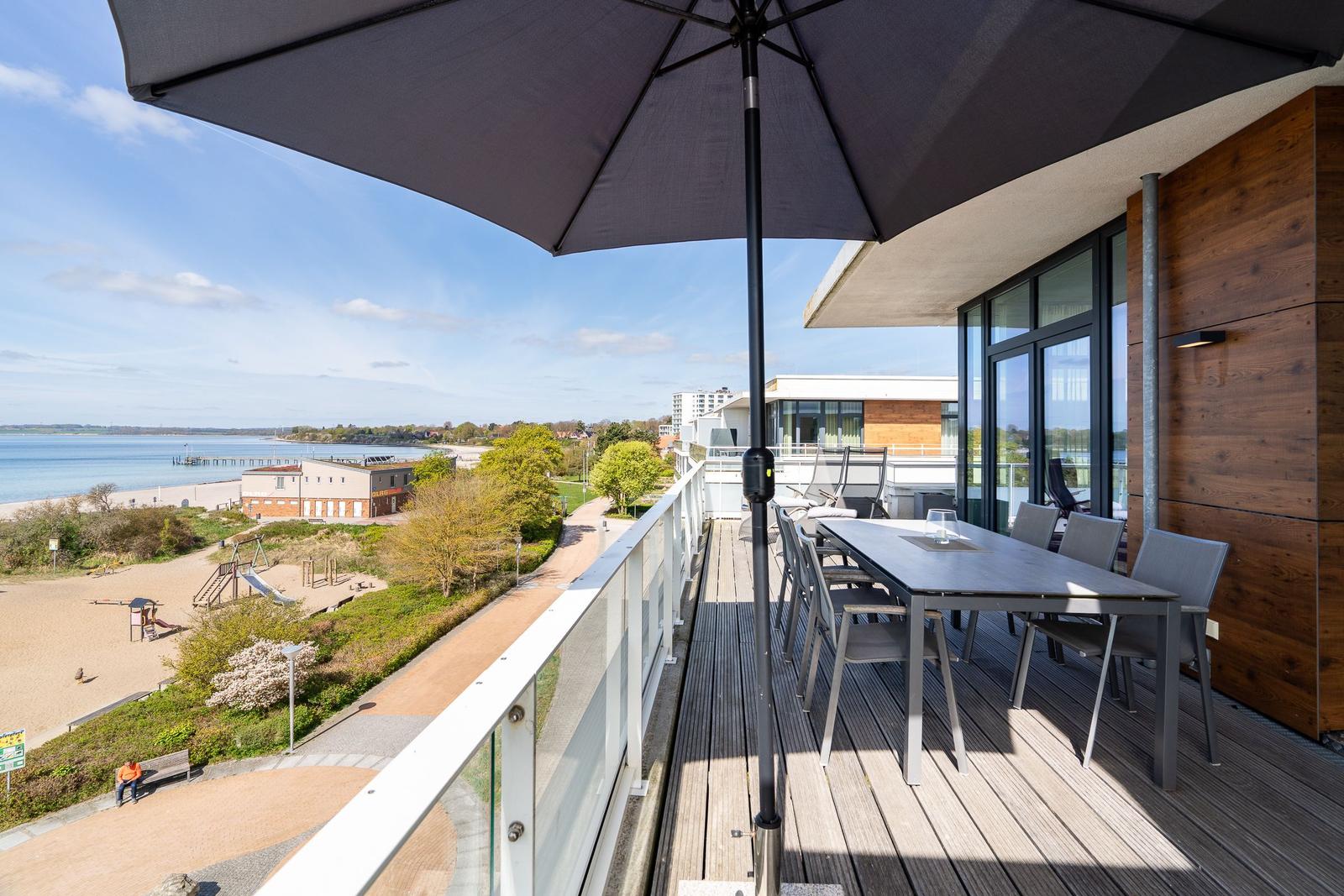 Terrasse mit Tisch, Stühlen und Sonnenschirm. Blick auf Strand und Wasser.