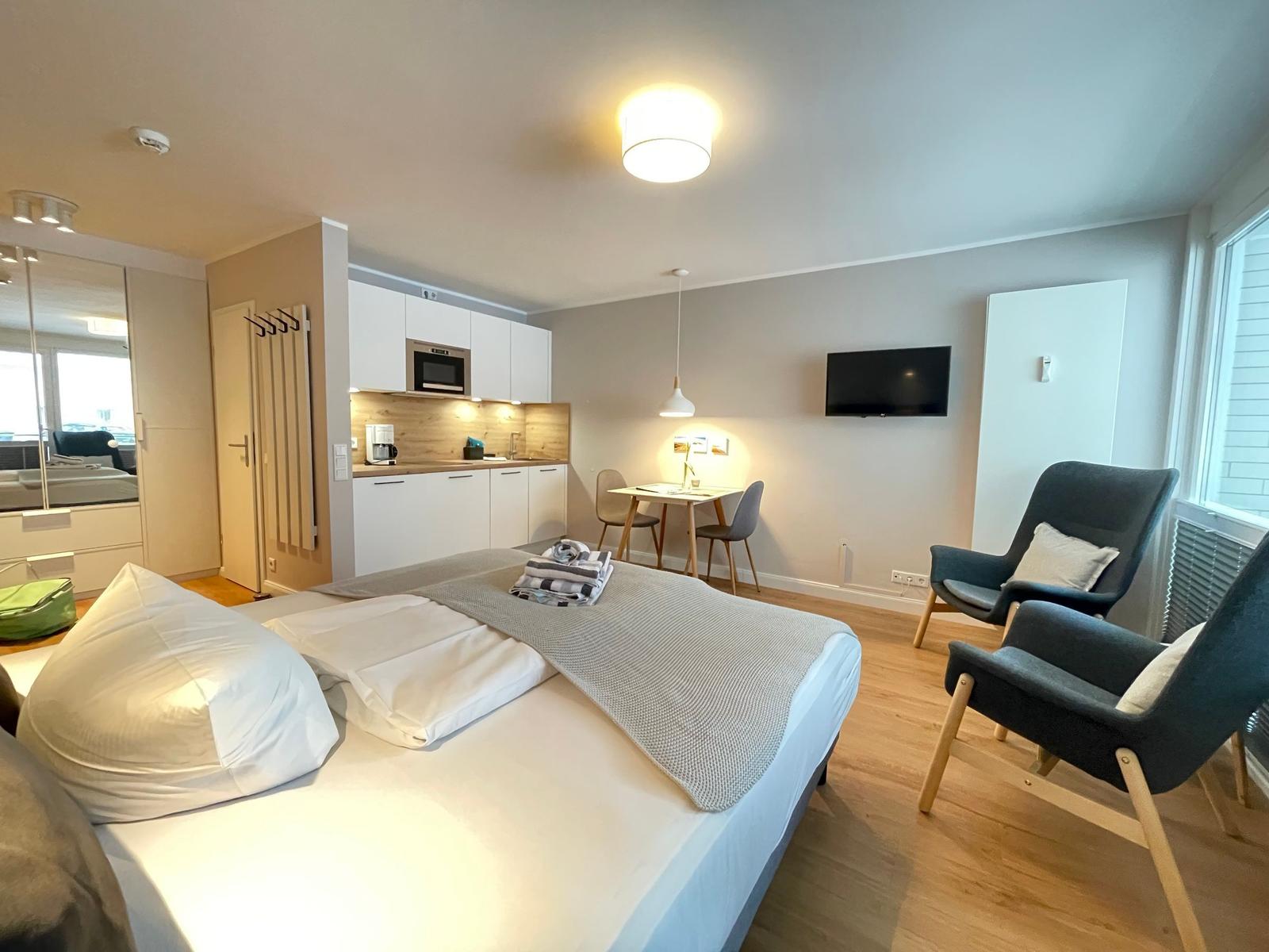 Schlafzimmer mit Bett, Küche und Sitzgelegenheiten. Wandfernseher und große Fenster.