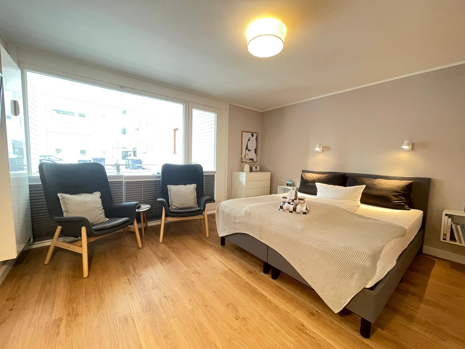 Schlafzimmer mit Bett, zwei Sesseln und großem Fenster.