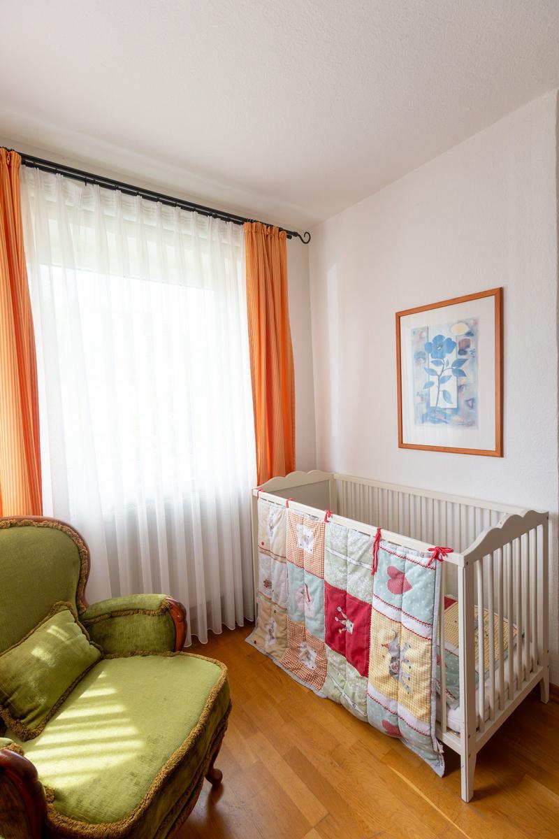 Ein Kinderzimmer mit einem weißen Kinderbett, einem grünen Sessel und orangefarbenen Vorhängen.