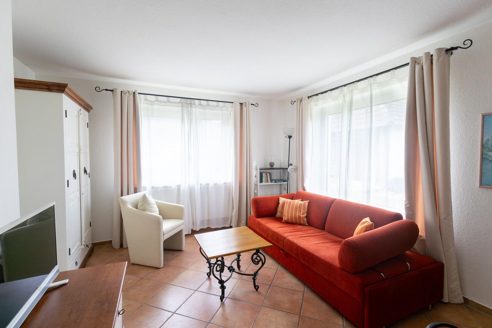 Wohnzimmer mit rotem Sofa, weißem Sessel und Holztisch. Fenster mit weißen Vorhängen.