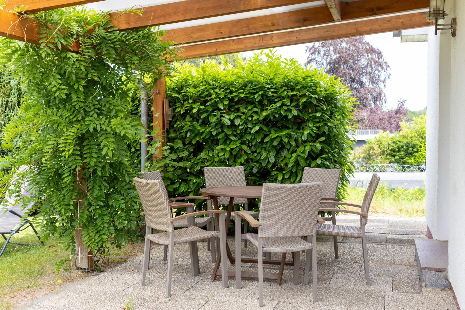 Terrasse mit Tisch und Stühlen unter einem Holzvordach. Grüne Hecke und Pflanzen im Hintergrund.