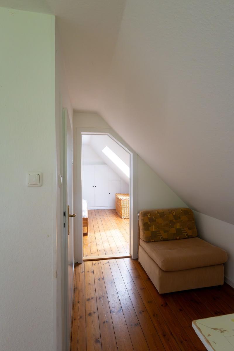 Dachgeschosszimmer mit Holzboden und Sitzgelegenheit. Durchgang zu Schlafzimmer mit Schrank und Korb.
