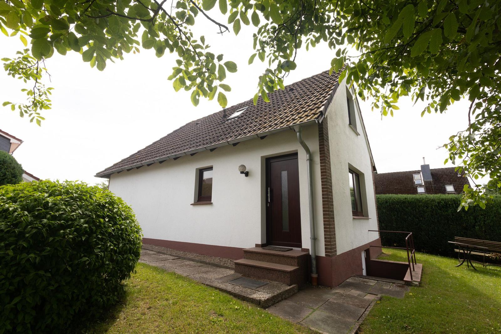 Ein kleines Haus mit weißer Fassade, braunem Dach und Vorgarten. Eingangstreppe und Fenster sind sichtbar.