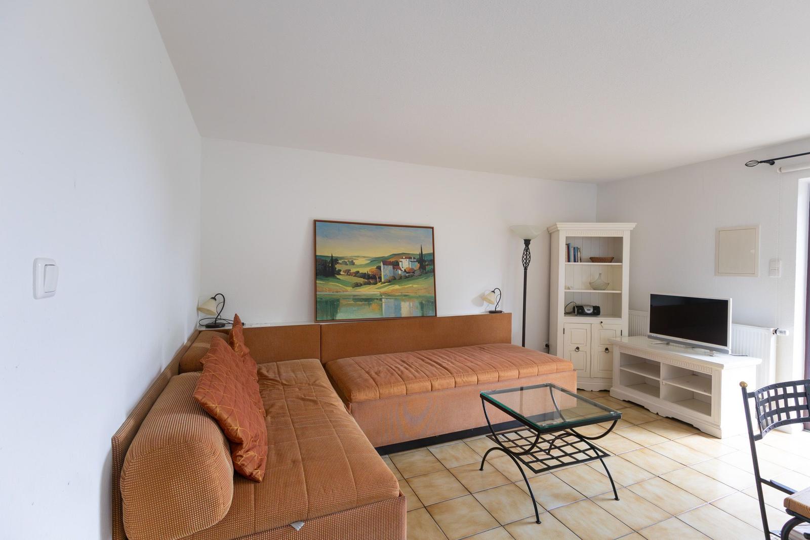 Wohnzimmer mit braunem Sofa, Fernseher und Glas-Tisch auf Fliesenboden.