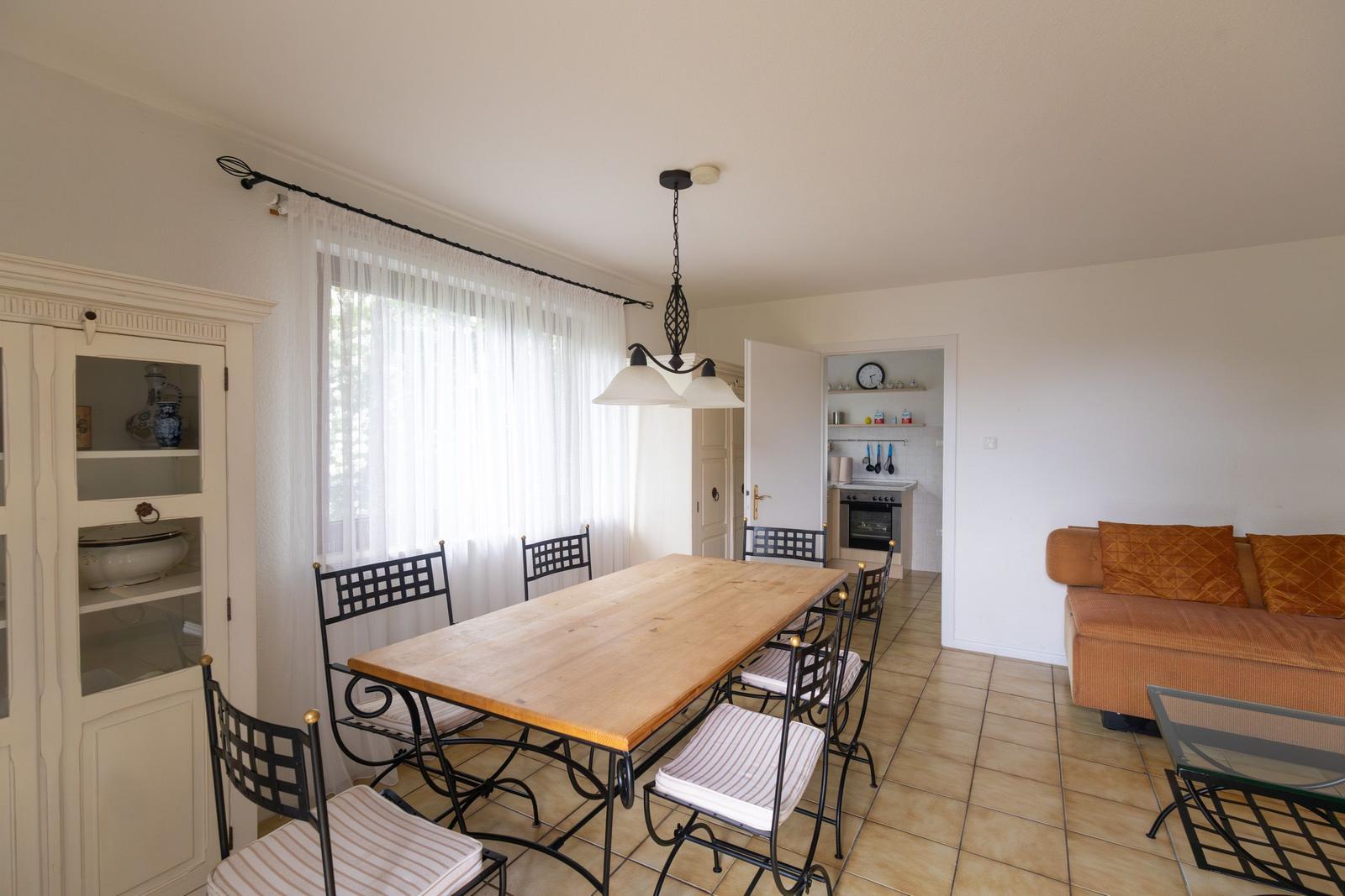 Essbereich mit Holztisch, Stühlen und Sofa im Wohnbereich. Küche im Hintergrund sichtbar.