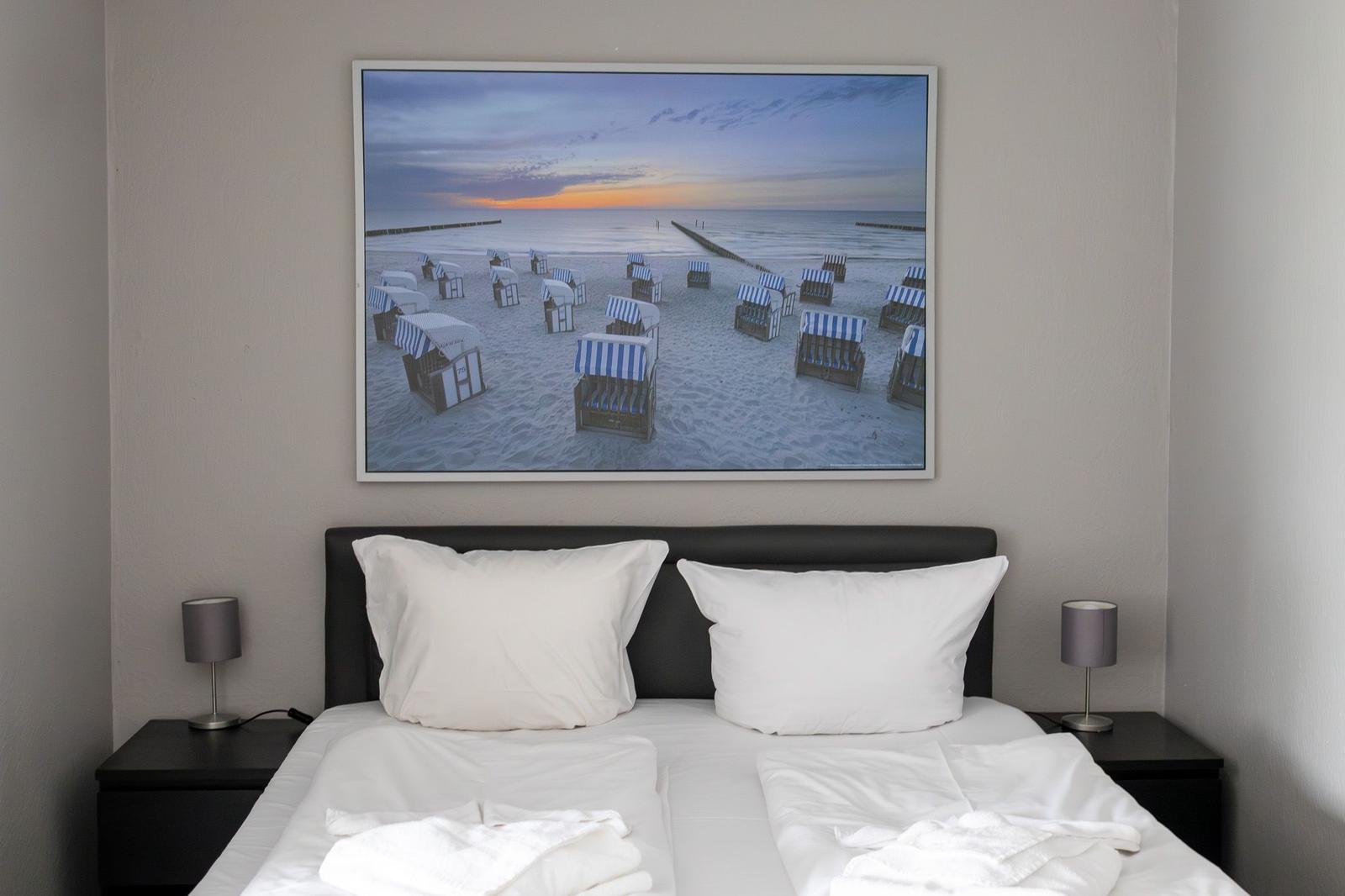 Schlafzimmer mit Bett, zwei Nachttischen und Wandbild mit Strandmotiv.
