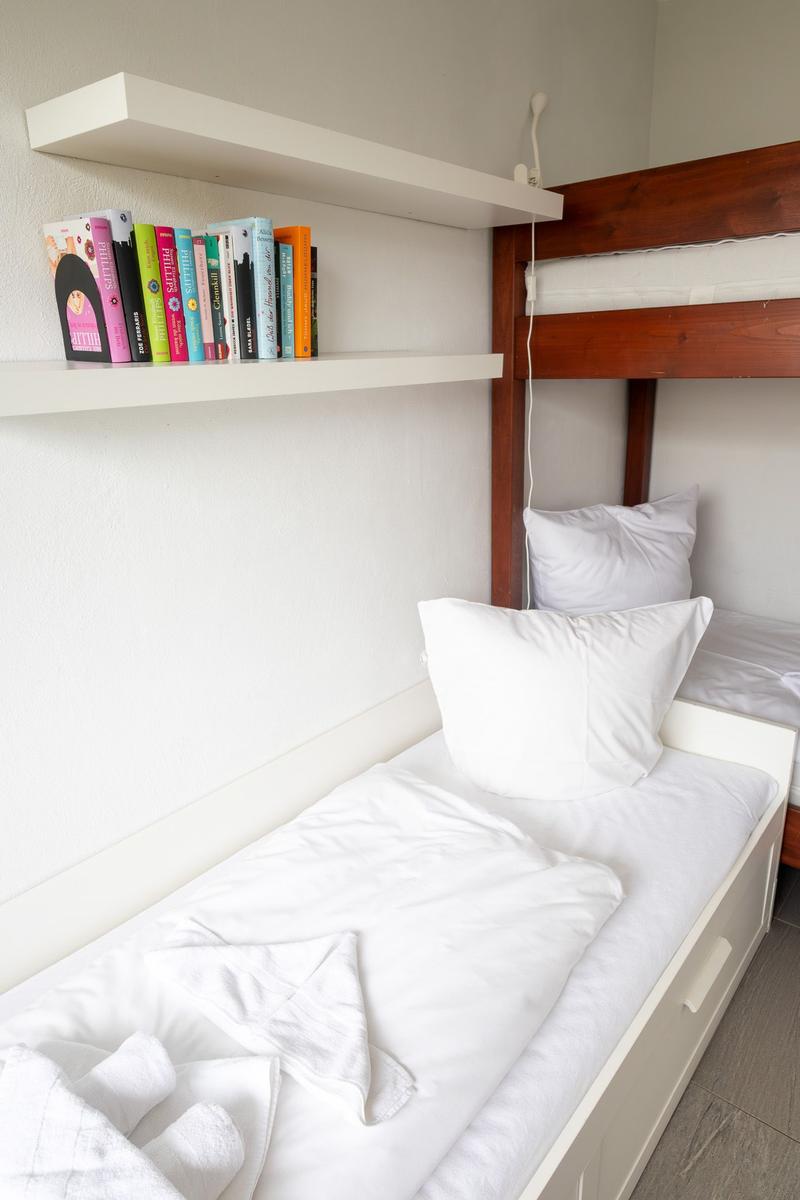 Ein Bett mit weißer Bettwäsche und einem Kissen. Über dem Bett hängt eine Stehlampe. Auf einem Regal stehen Bücher.