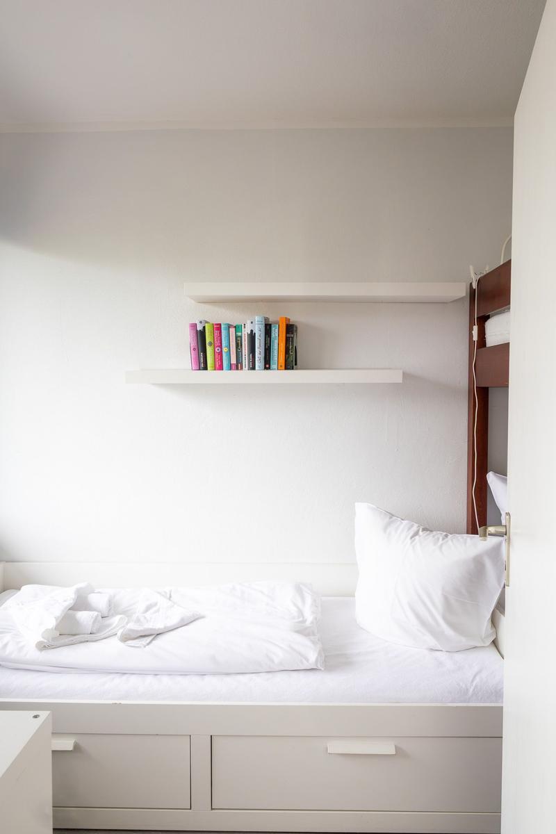 Ein Bett mit weißen Laken und Kissen. An der Wand hängen zwei weiße Regale mit Büchern.