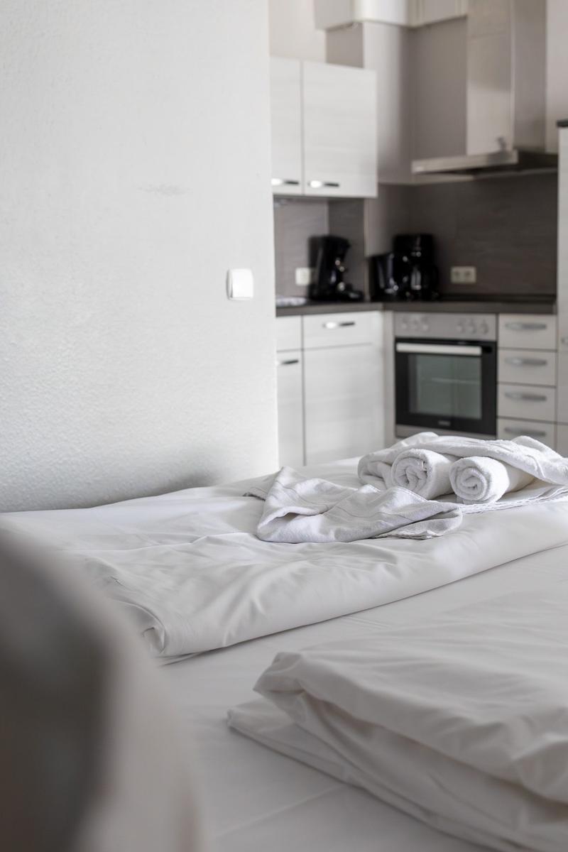 Ein Bett mit weißen Laken und Handtüchern. Im Hintergrund ist eine Küche mit weißen Schränken.