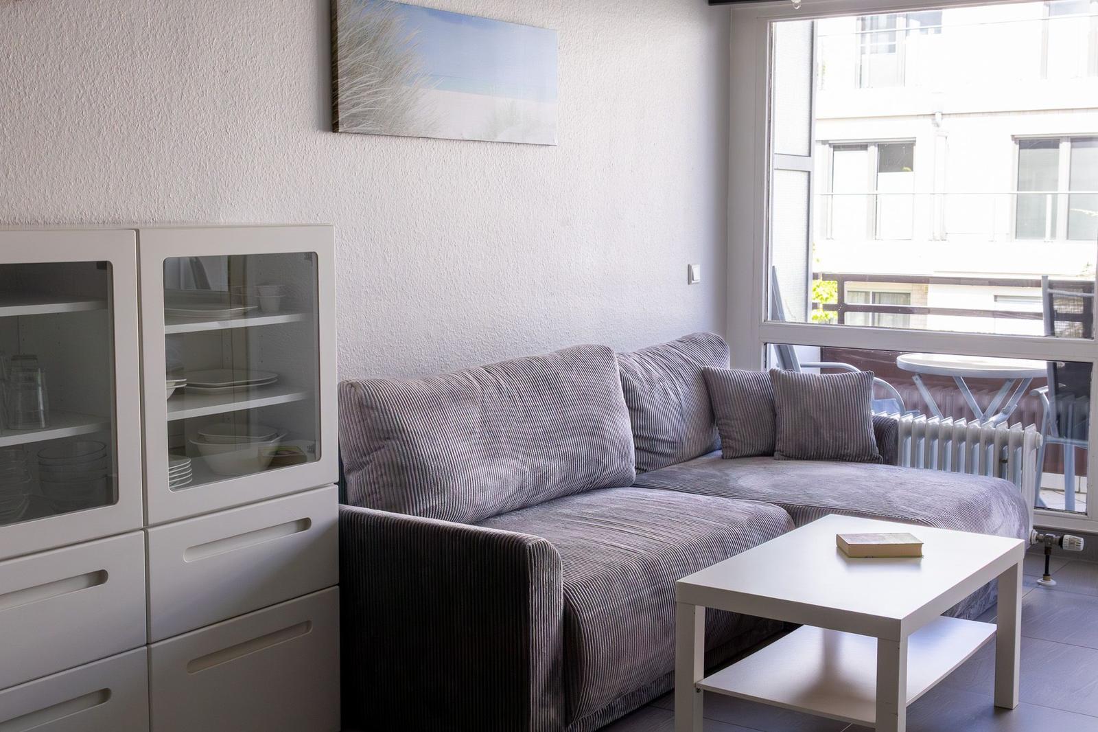 Ein Wohnzimmer mit grauem Sofa, weißem Couchtisch und Glasvitrine. Durch Fenster ist Balkon sichtbar.
