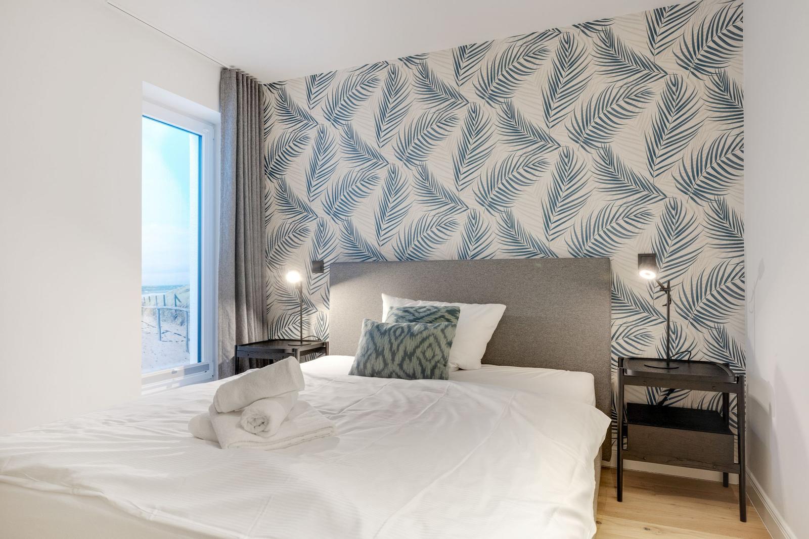 Schlafzimmer mit Bett, Fenster und Palmenwandtapete.