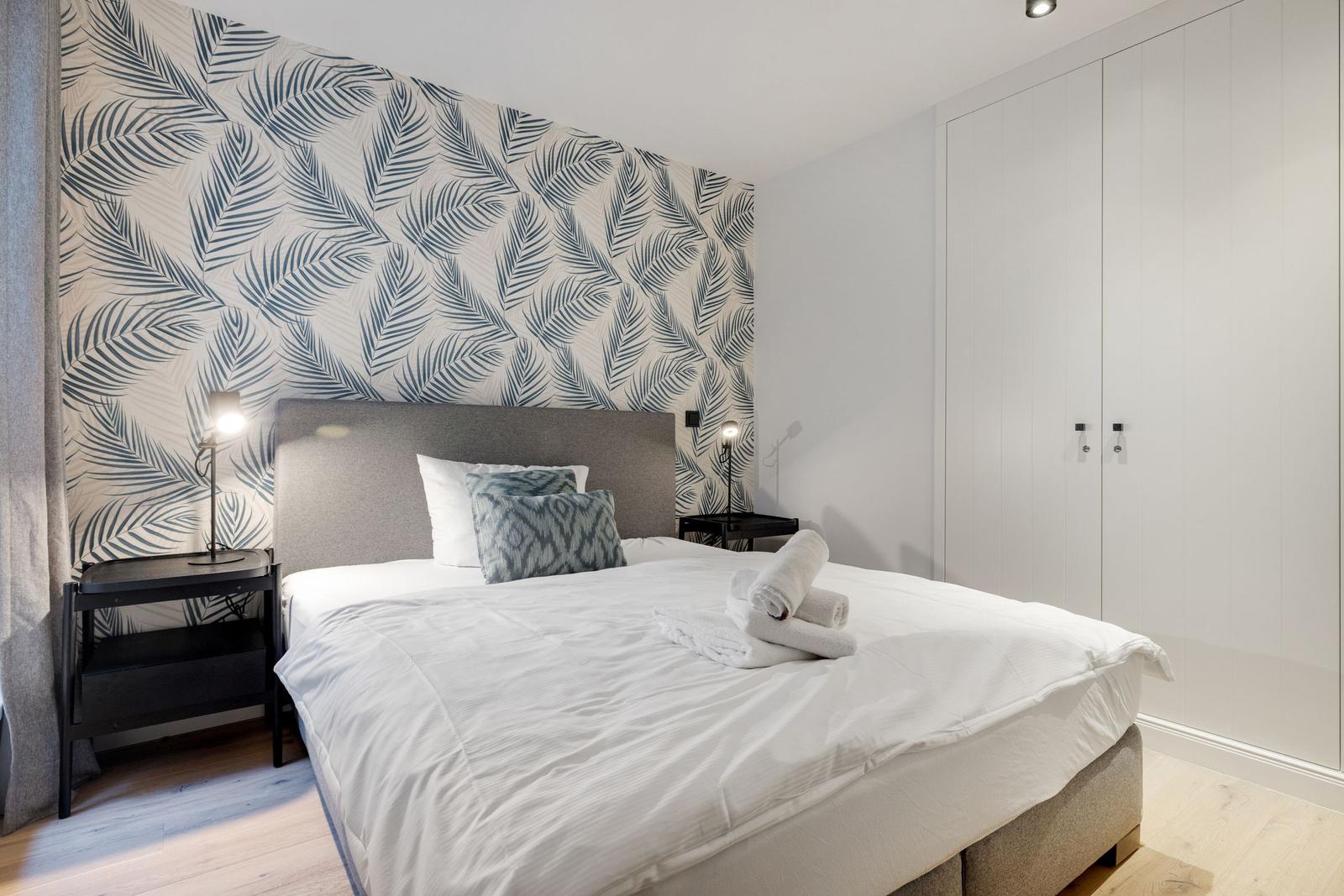 Schlafzimmer mit Bett, Nachttischen und Tapete mit Blättern.