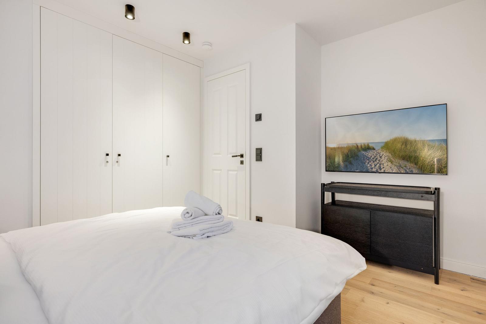 Schlafzimmer mit weißem Bett, Kleiderschrank und Fernseher an der Wand.