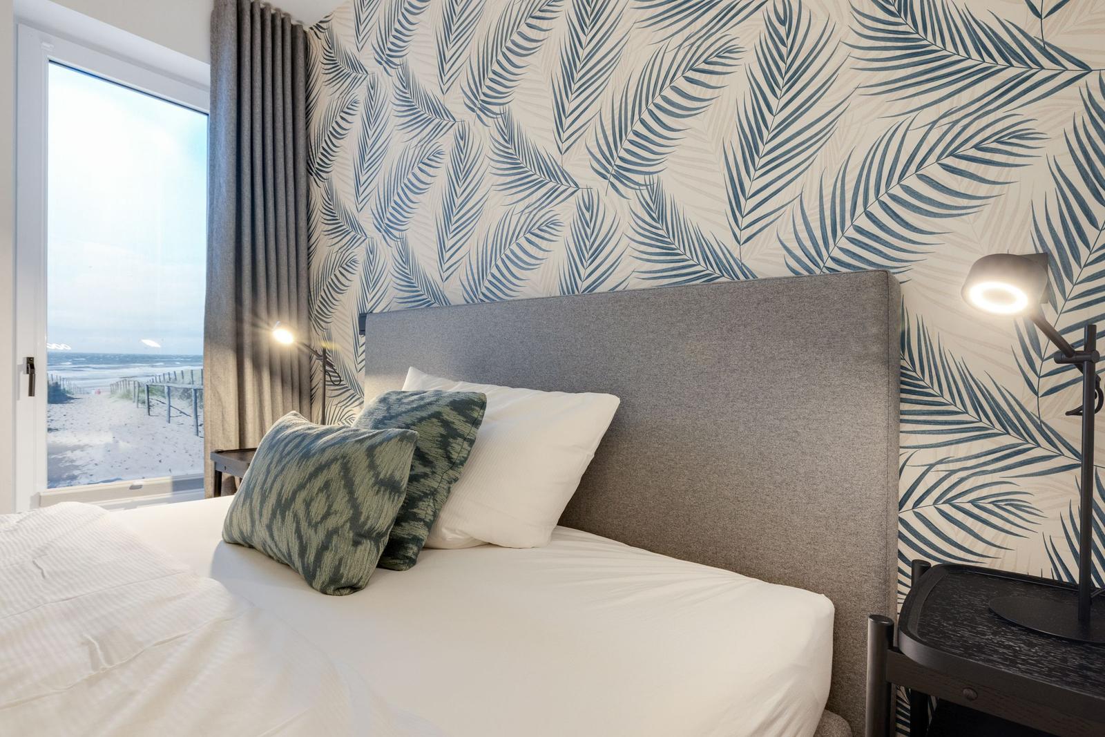 Schlafzimmer mit Blick auf Strand, Palmen Tapete und Nachttisch mit Lampe.