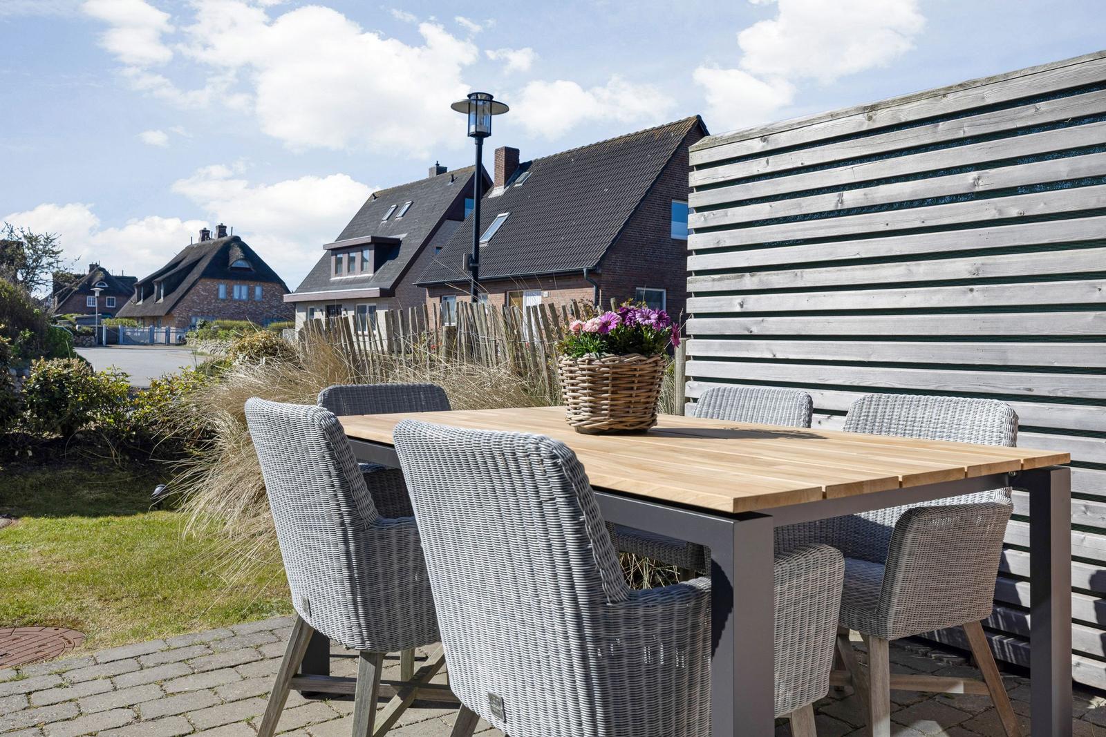 Terrasse mit Holztisch und sechs Stühlen, Blumenkorb, Holz-Zaun und Hintergrundhäuser.