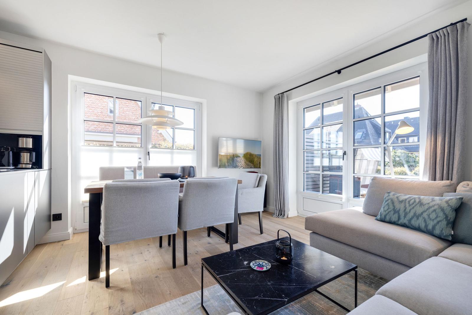 Wohn- und Essbereich mit Sofa, Tisch und Stühlen. Große Fenster und helles Holzfußboden.