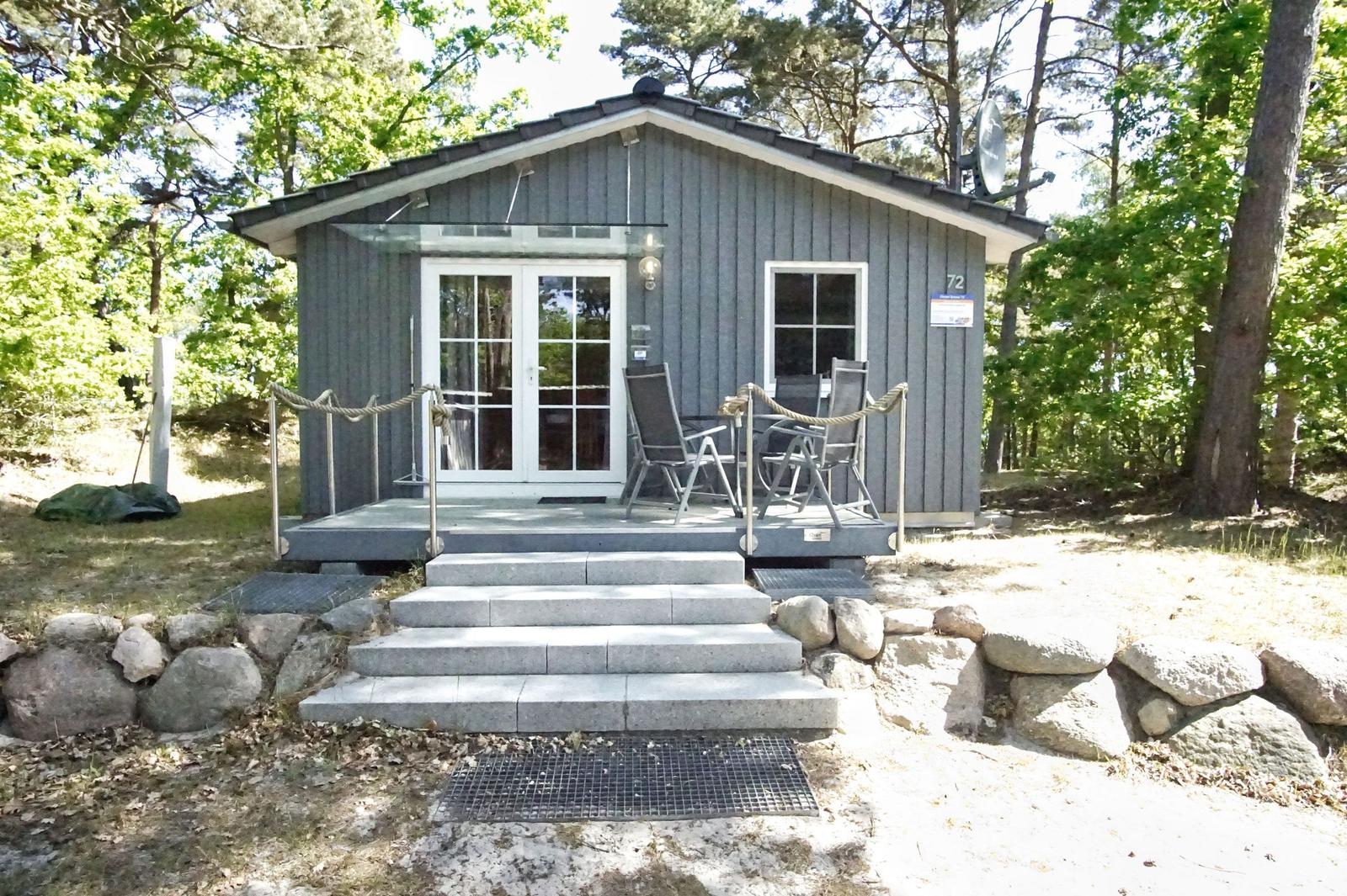 Ein graues Holzhaus mit Terrasse, Stufen und Garten. Umgeben von Bäumen.