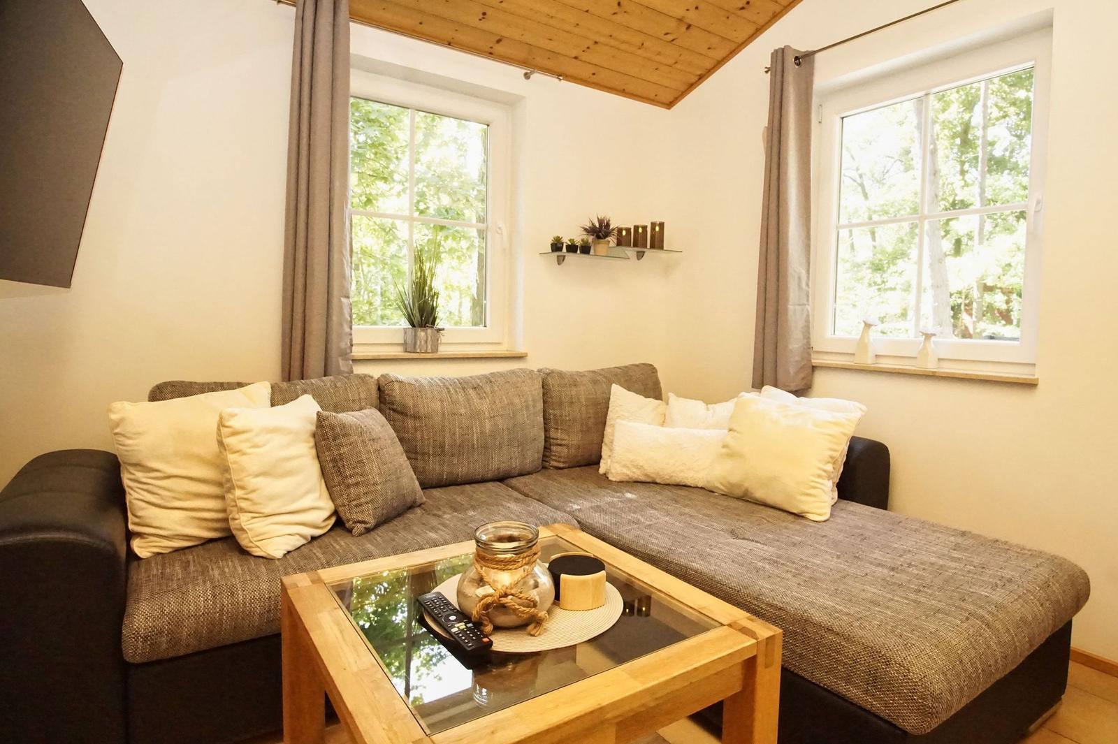 Ein Wohnzimmer mit Ecksofa, Glas-Tisch und Fenstern mit Blick auf Bäume.