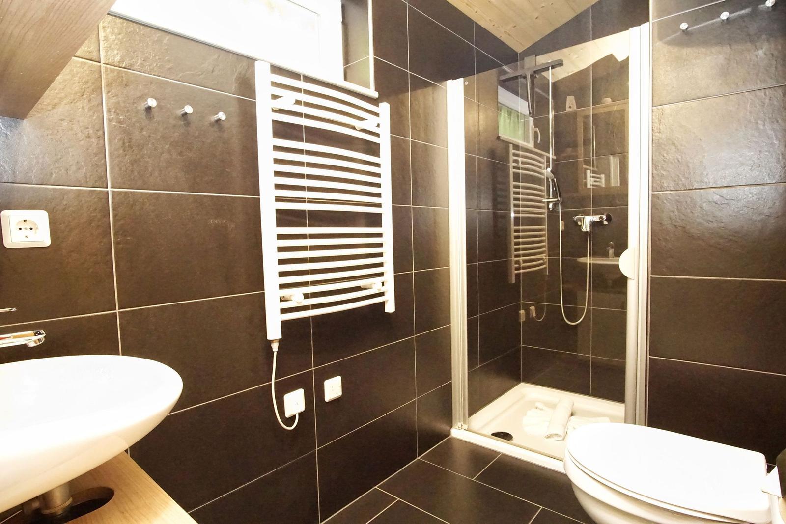 Badezimmer mit Dusche, Waschbecken und Toilette. Wände und Boden sind mit dunklen Fliesen verkleidet.